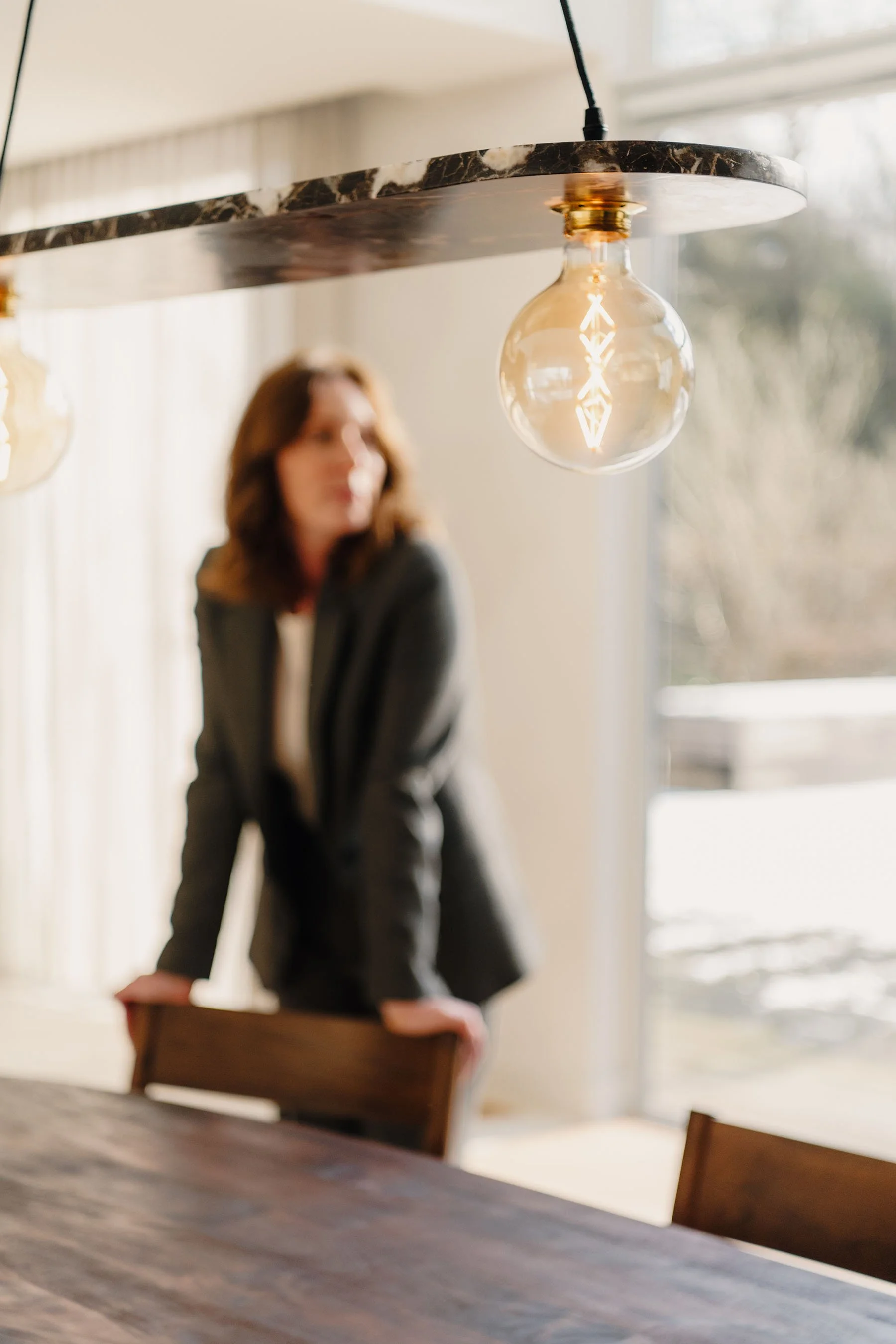 Een vrouw in een grijze blazer leunt met haar handen op een houten tafel in een lichte kamer met grote ramen.