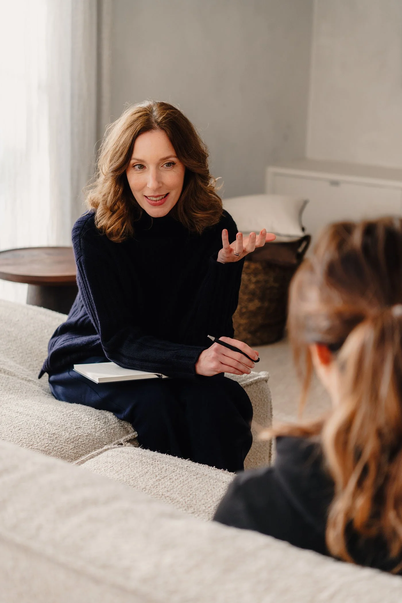 Twee vrouwen hebben een gesprek in een woonkamer, de ene vrouw zit op een bank en de andere zit met haar rug naar de camera.
