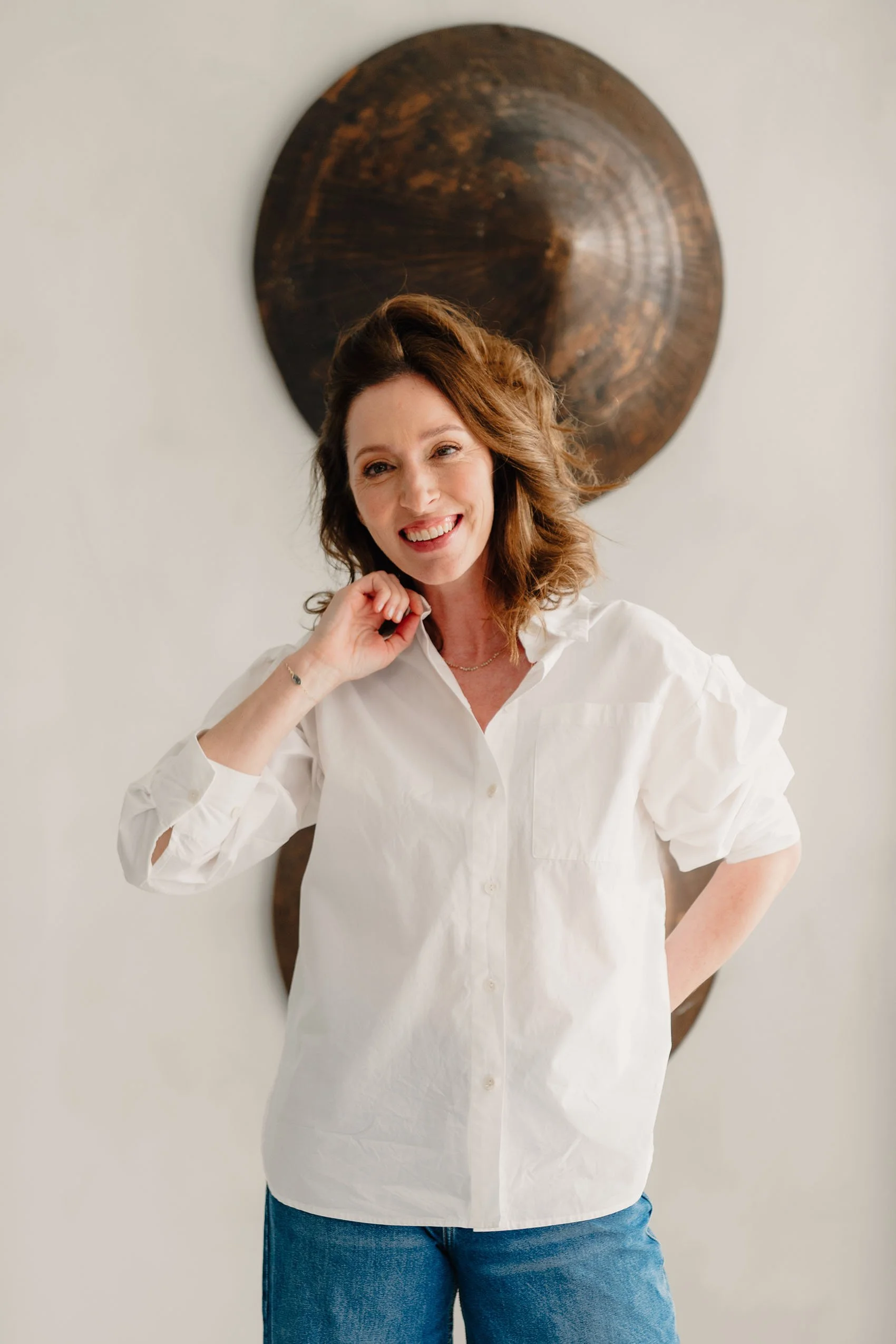 Vrouw met bruin haar in een witte blouse en blauwe jeans lachend voor een witte muur met een decoratief houten wandkunstwerk.