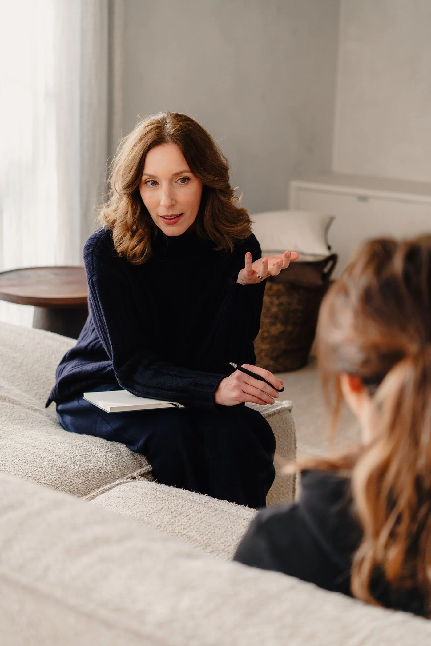 Vrouw in gesprek met therapeut in woonkamer, zit op bank met notitieboekje.