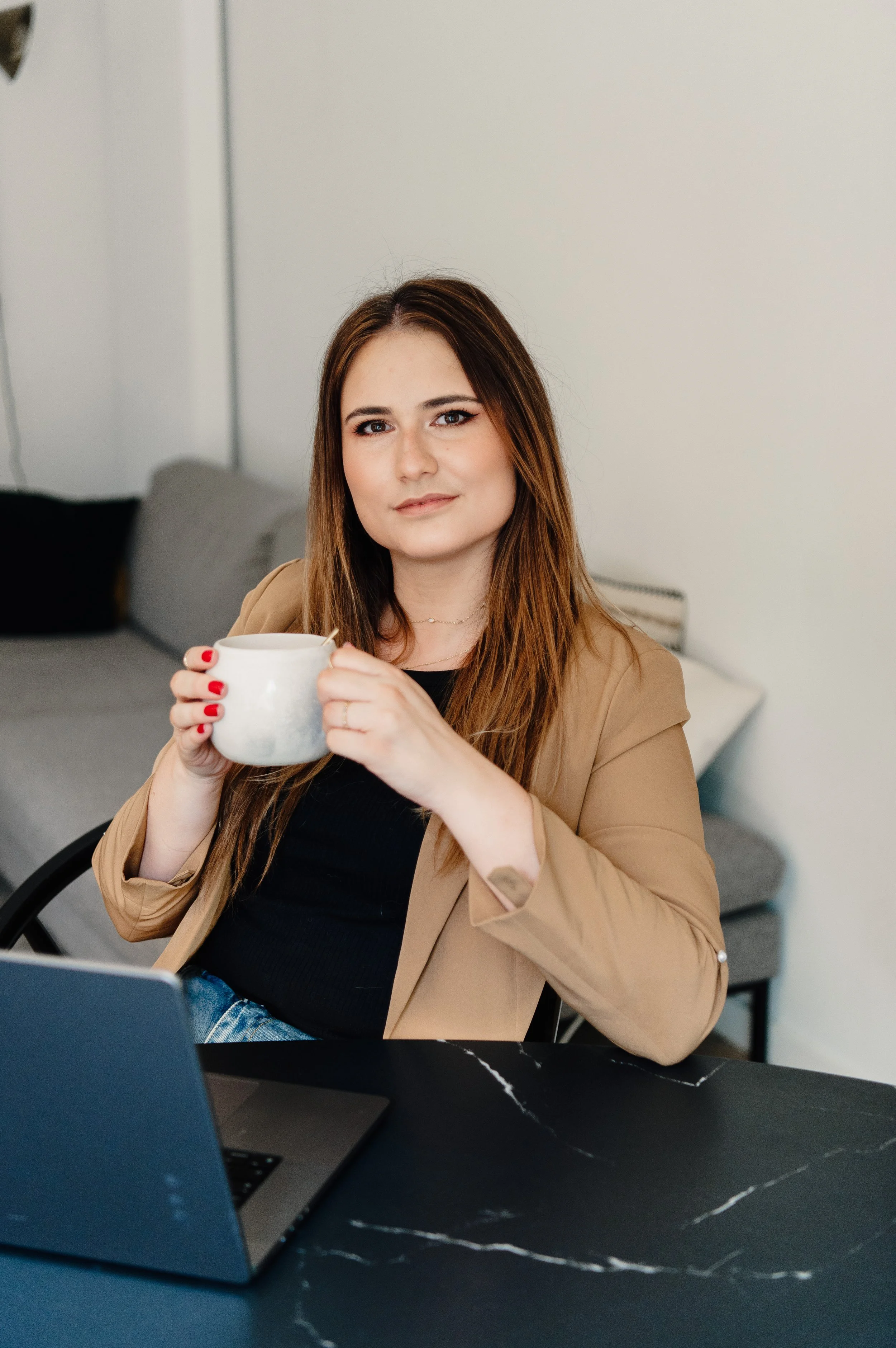 Een vrouw met lang bruin haar houdt een witte mok vast terwijl ze achter een laptop zit in een woonkamer.