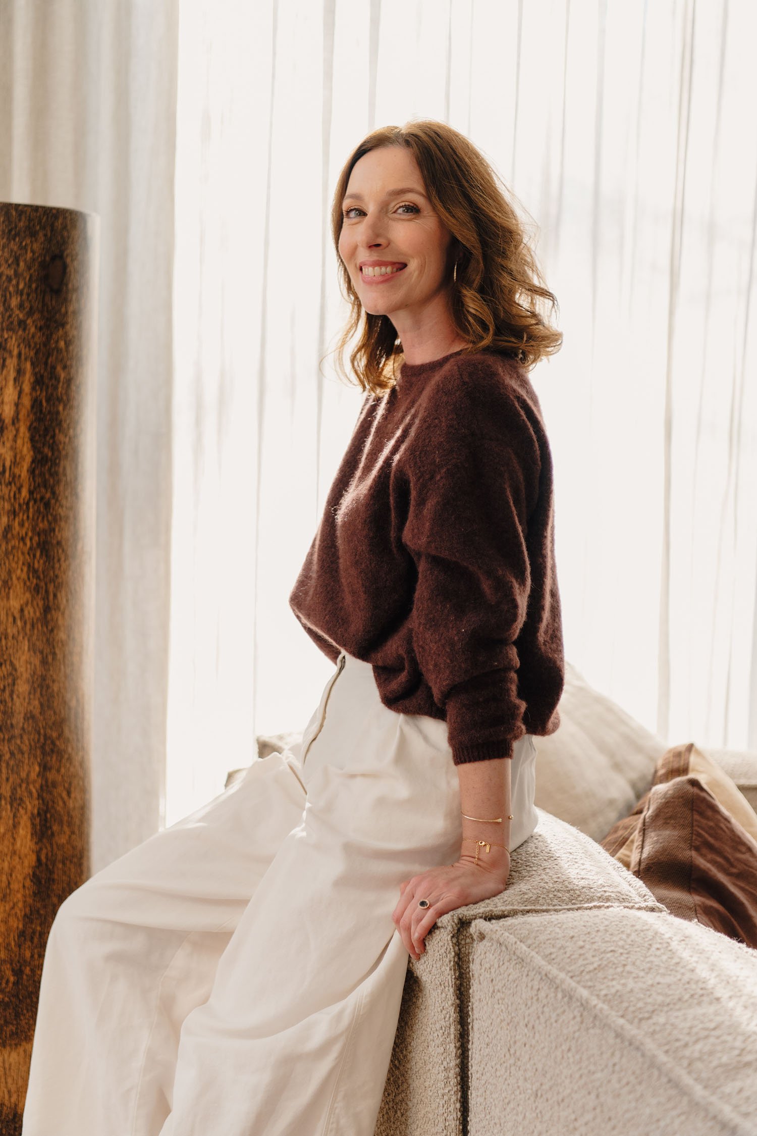 Vrouw met bruin haar die op een bank leunt, binnen, gekleed in een bruine trui en witte broek, lachend en naar de camera kijkend.