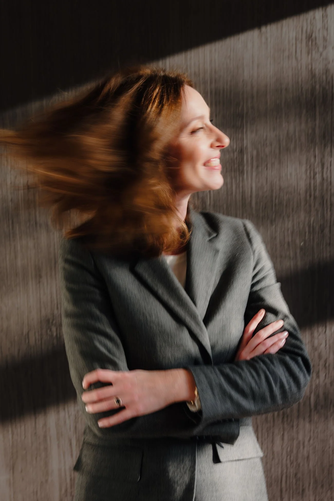 Portret van een lachende vrouw met rood haar in zakelijk pak, haar haar beweegt door beweging of wind, tegen een houten achtergrond.