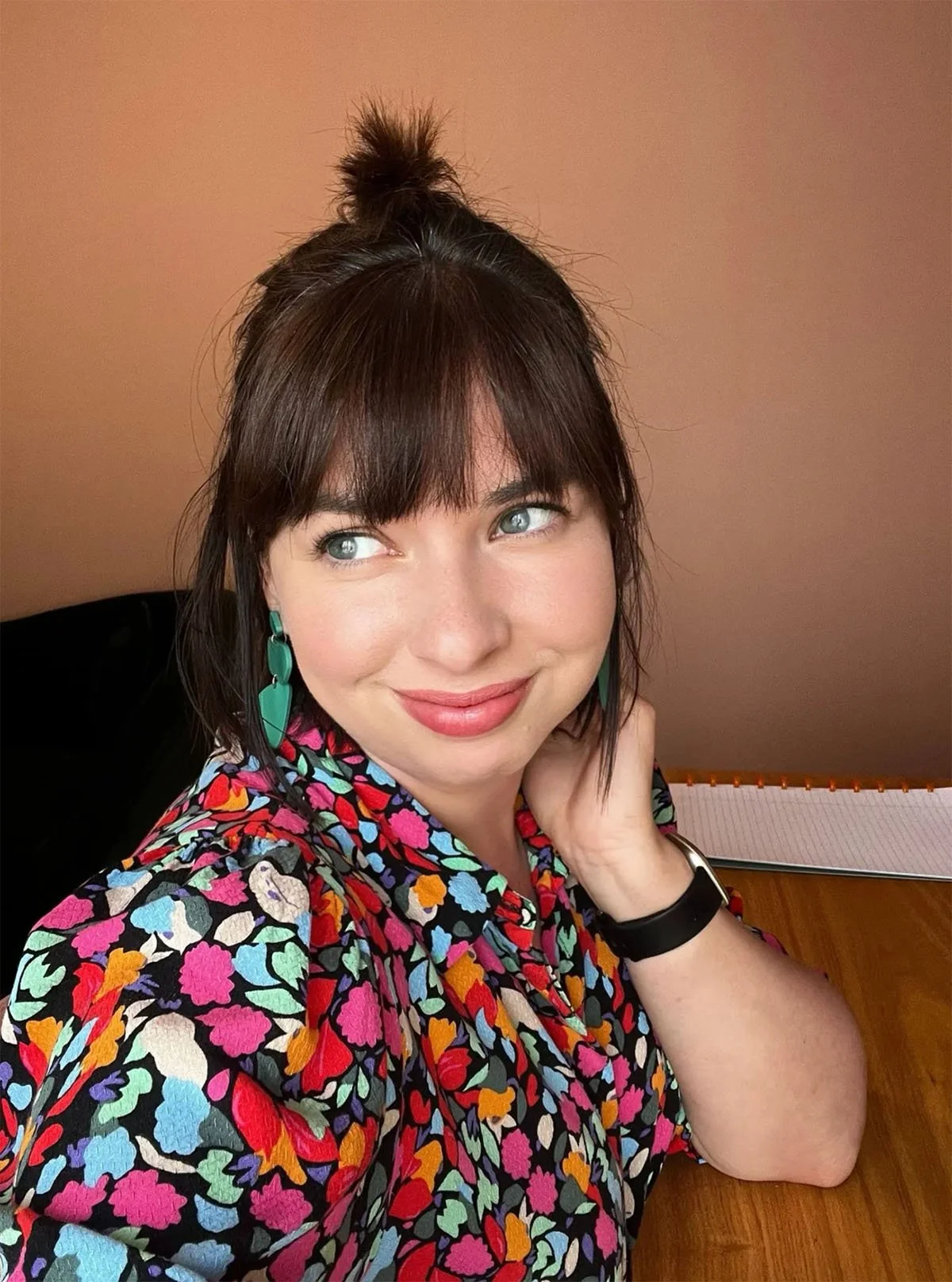 Een vrouw met bruin haar en een hoge paardenstaart, gekleed in een kleurrijk bloemenpatroon shirt, glimlachend en naar links kijkend, zittend aan een houten tafel met een notitieboek op de achtergrond.