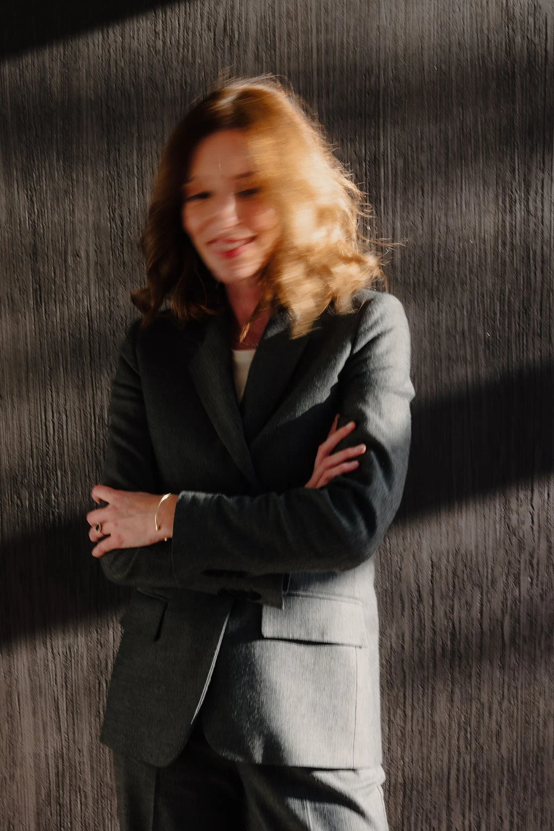 Een vrouw met schouderlange, lichtbruin haar, gekleed in een grijs pak en een witte blouse, staat met haar armen crossed voor een donkere, houten muur.