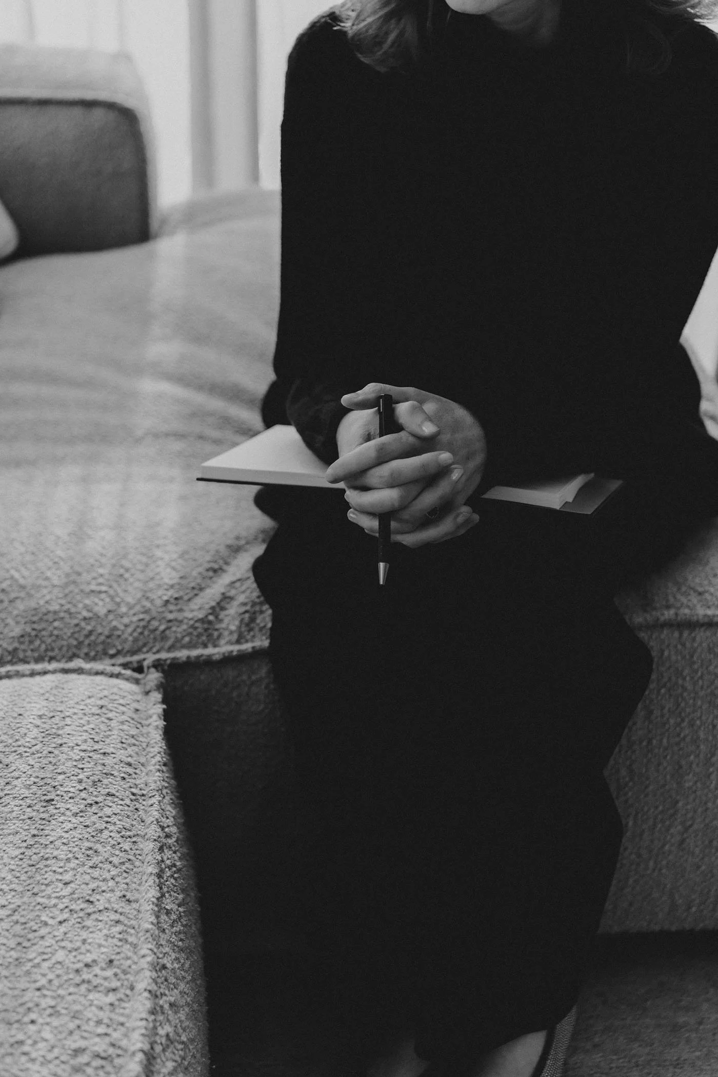 Een persoon zit op een bank, gekleed in zwart, met de handen geklemd om een pen en een notitieboekje.