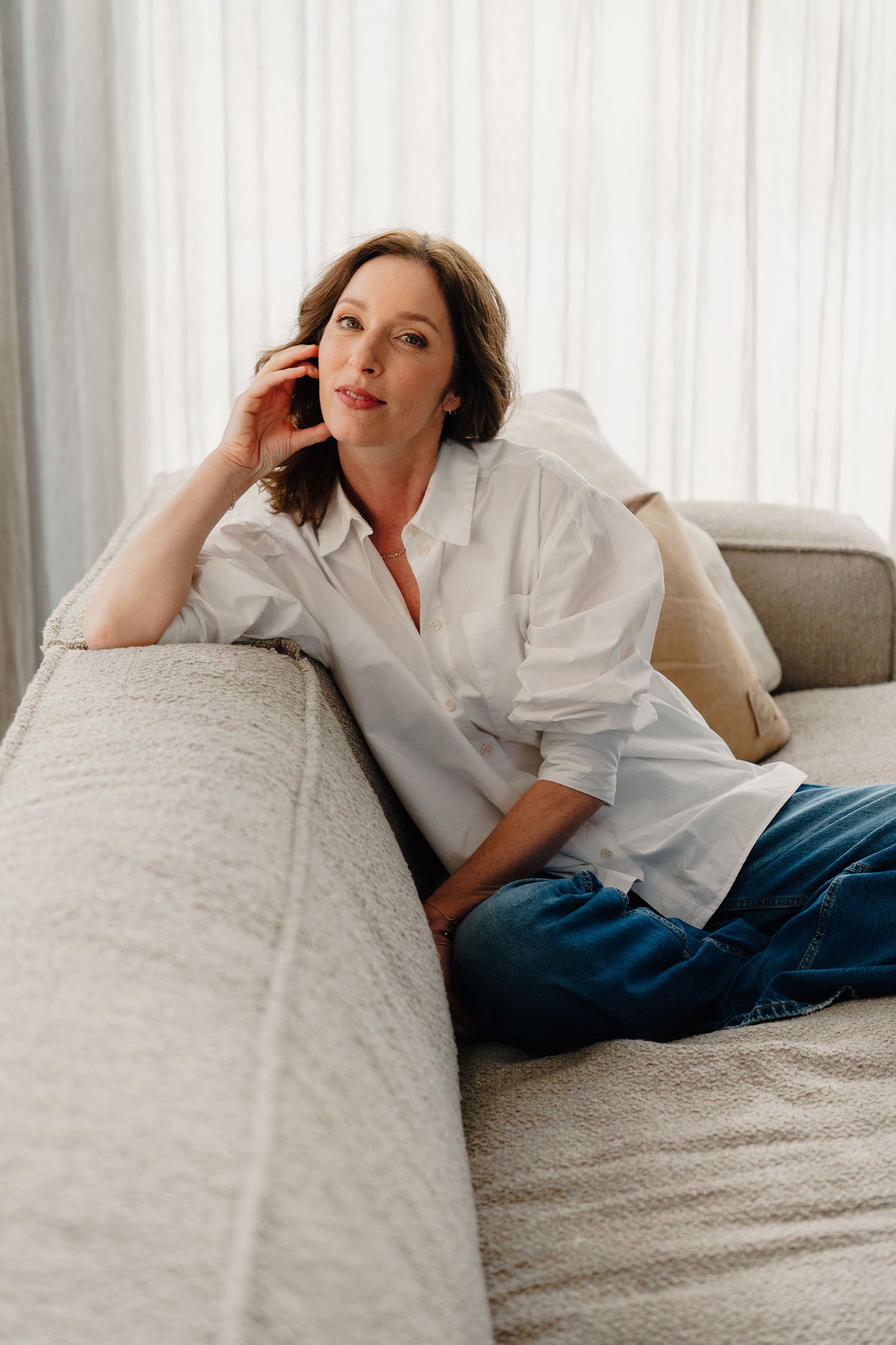 Een vrouw ligt ontspannen op een beige bank met kussens, gekleed in een witte blouse en blauwe jeans, voor een raam met lichte gordijnen.