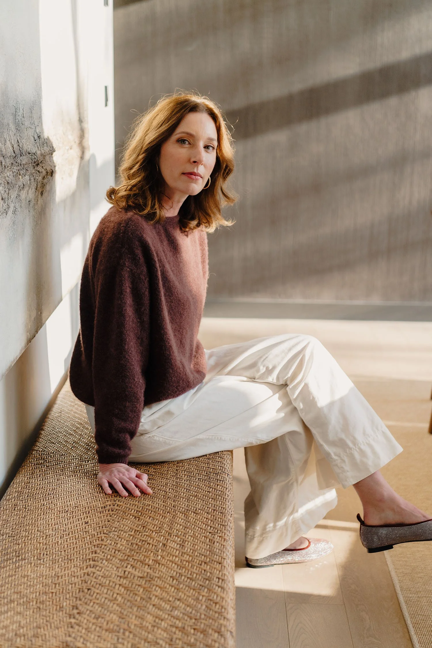 Een vrouw zit op een bankje tegen een muur, kijkt naar de camera, draagt een bruin sweater en witte broek, met sierlijke schoenen.