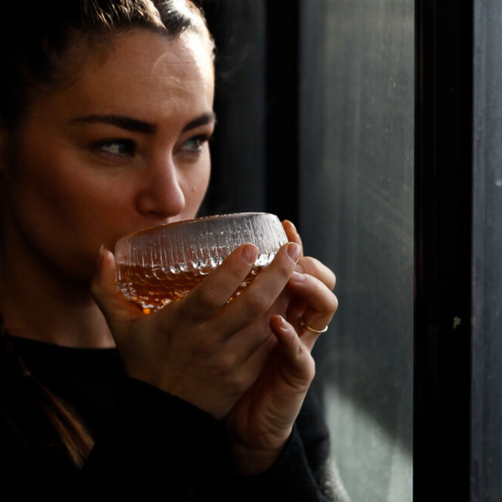 A woman holding a glass cup of vegan broth near a window, looking outside.