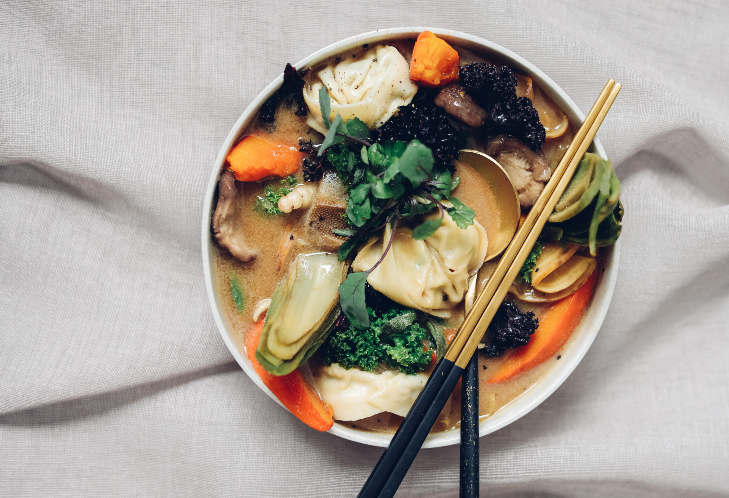 A bowl of Asian-style noodle soup with vegetables, dumplings, herbs, and chopsticks on a white fabric background.