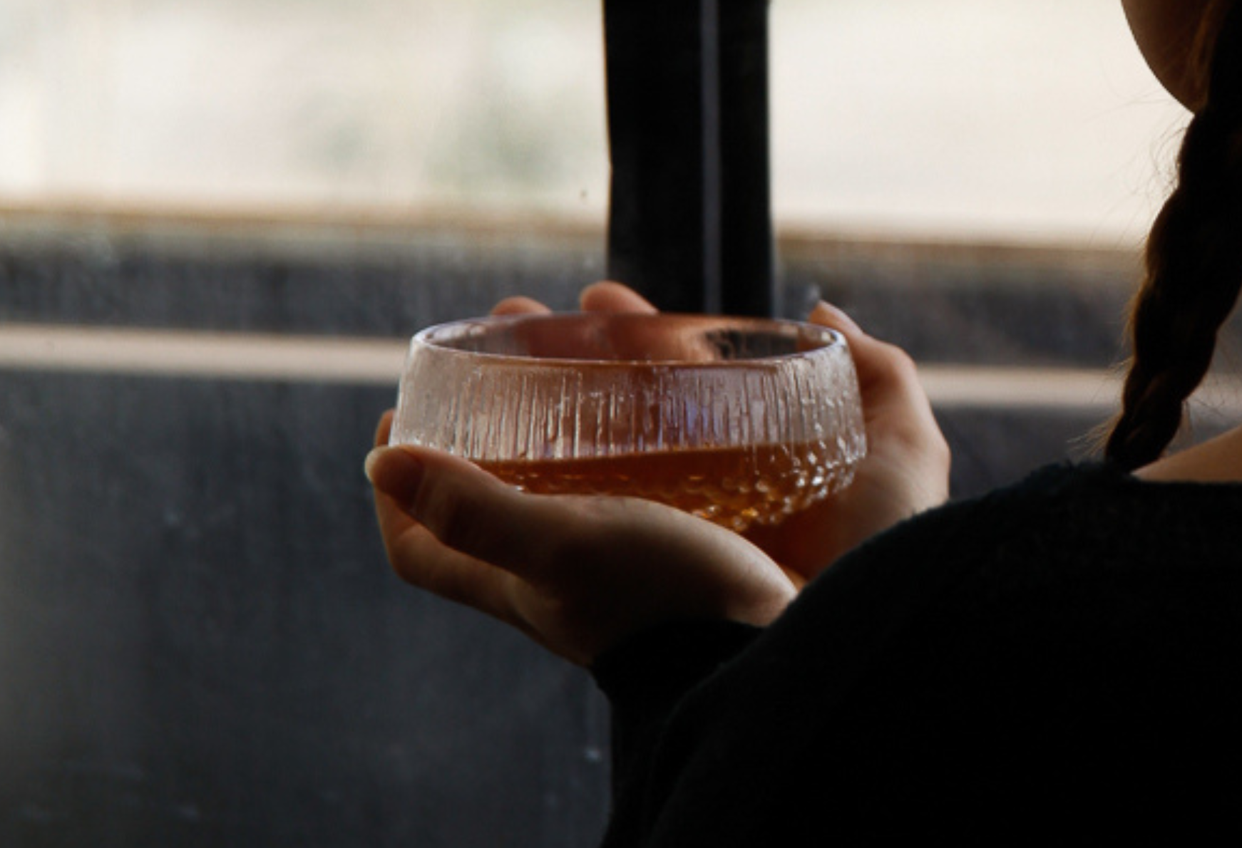 Person holding a glass bowl with broth, near a window.
