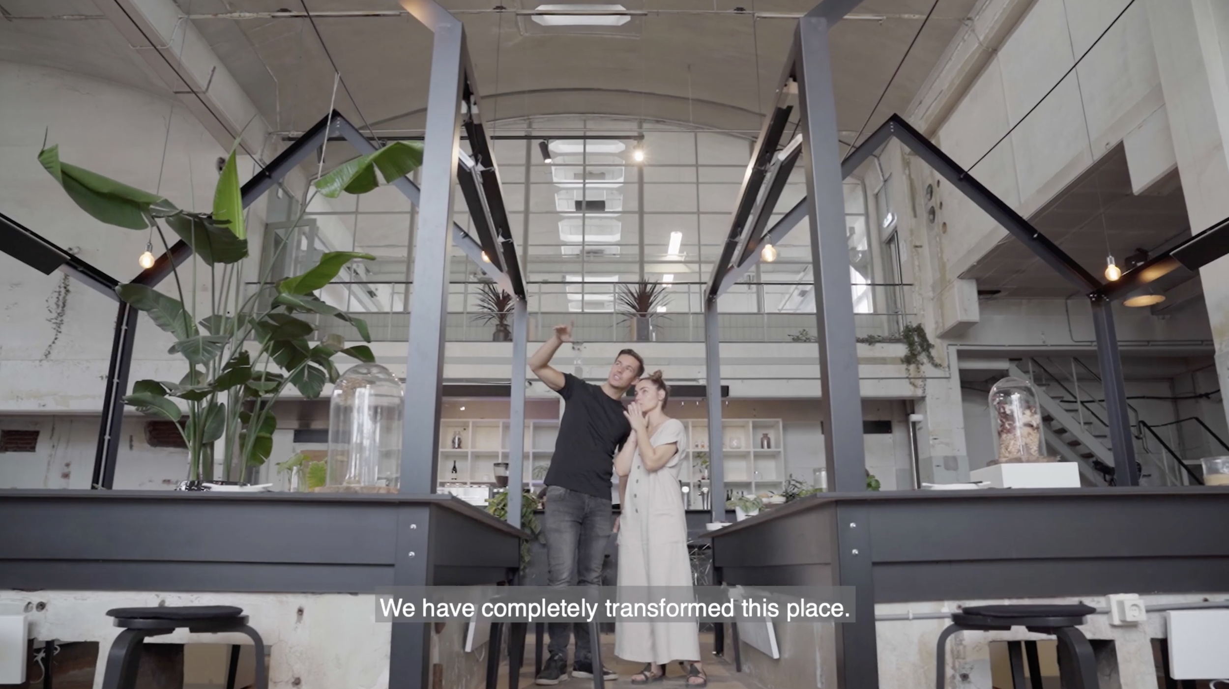 A man and woman standing in a modern, renovated industrial-style indoor space, looking at something while the man points upward, with subtitle 'We have completely transformed this place.'