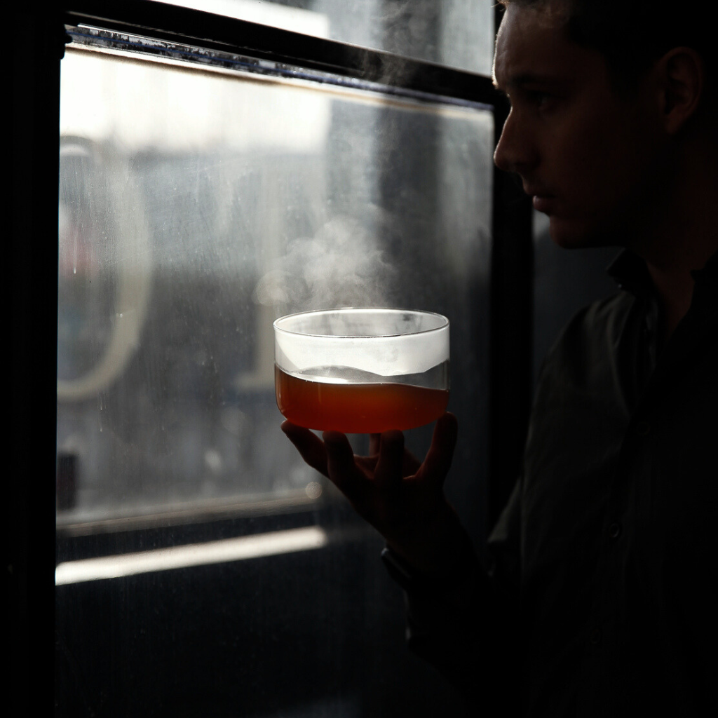 A man in silhouette holding a steaming bowl of amber-colored broth near a window, with steam rising from the drink.