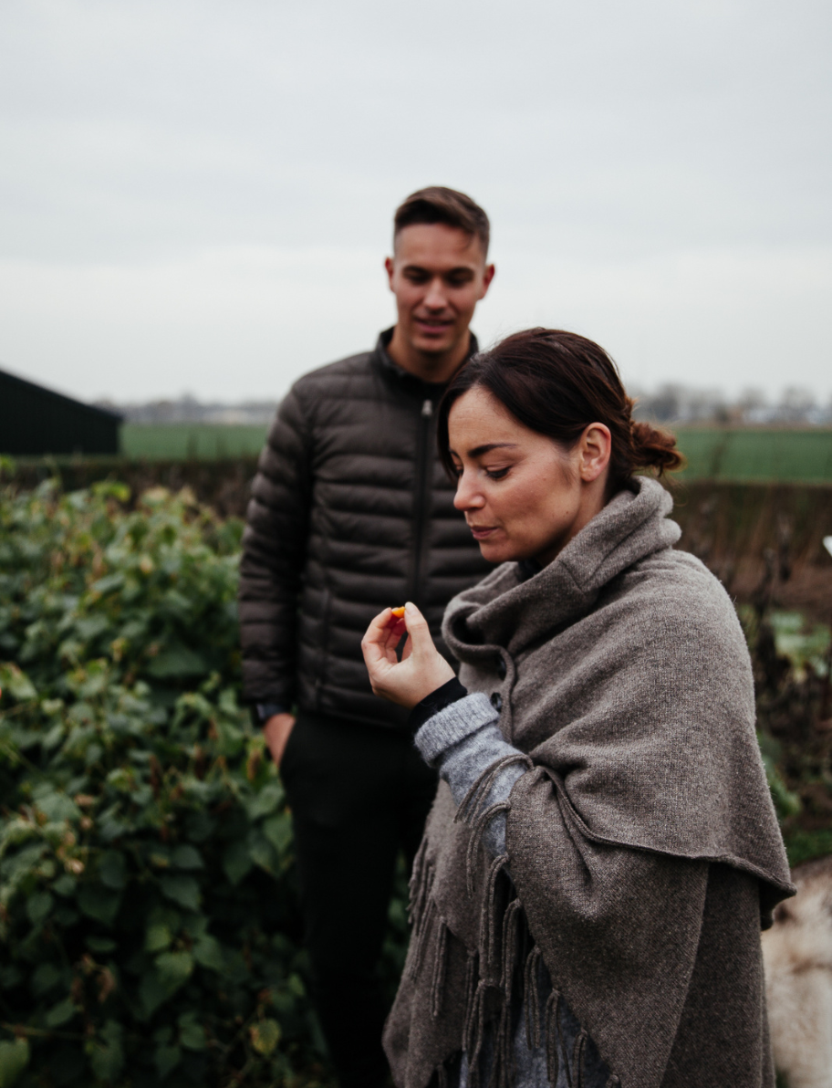 A woman wearing a gray shawl holding a small object, with a man in a black puffer jacket standing behind her in an outdoor setting with green bushes and cloudy sky.