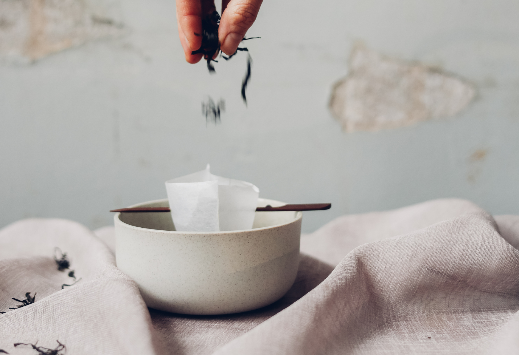 A hand dropping dried tea leaves into a bowl. The bowl contains a folded paper filter resting on a pair of chopsticks, with a soft pink cloth underneath. A textured wall is in the background.