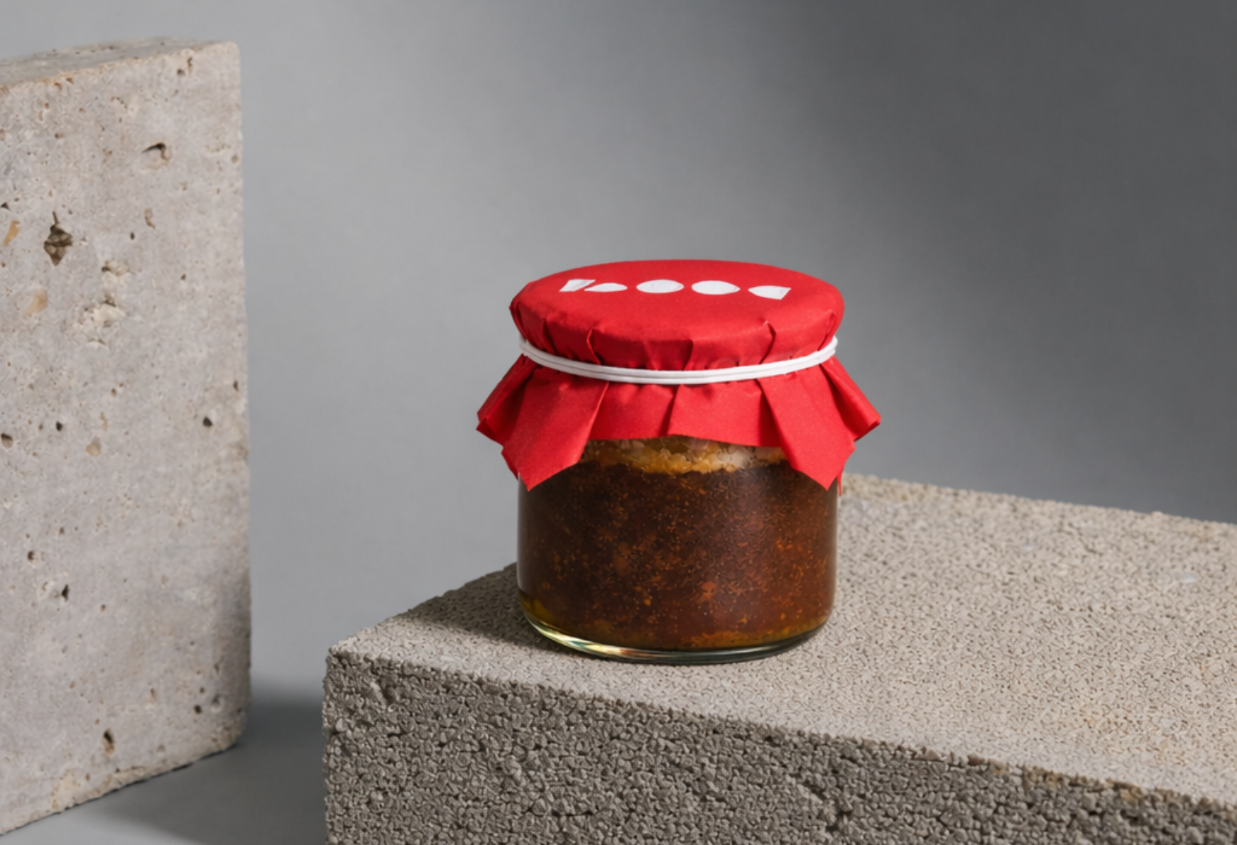 A jar of homemade vegan mineral broth with a red paper cover and a white rubber band, placed on a textured concrete block against a gray background.