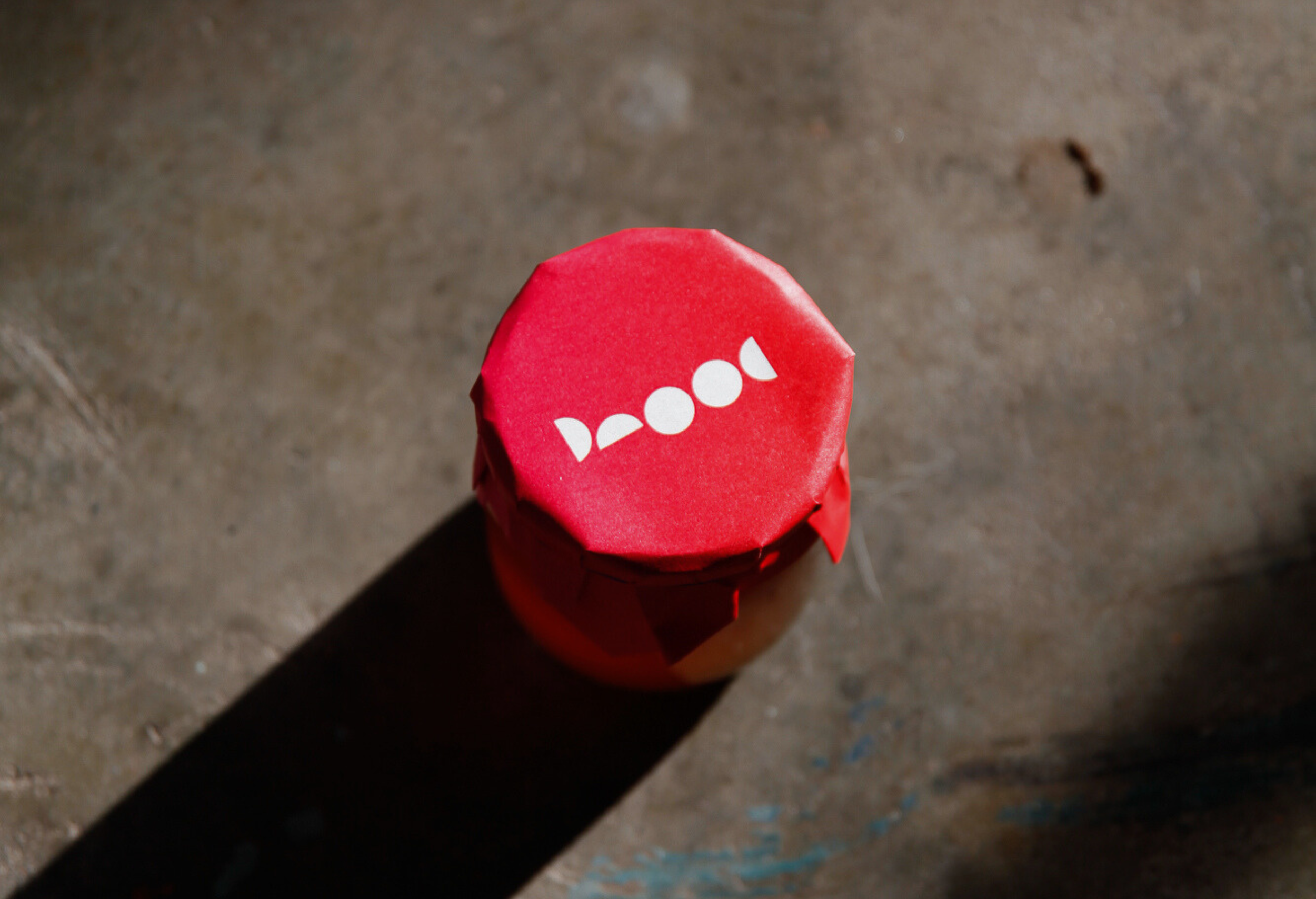 A jar covered with red paper and white stencil letters that spell out 'Phood' on a concrete surface, casting a shadow to the left.