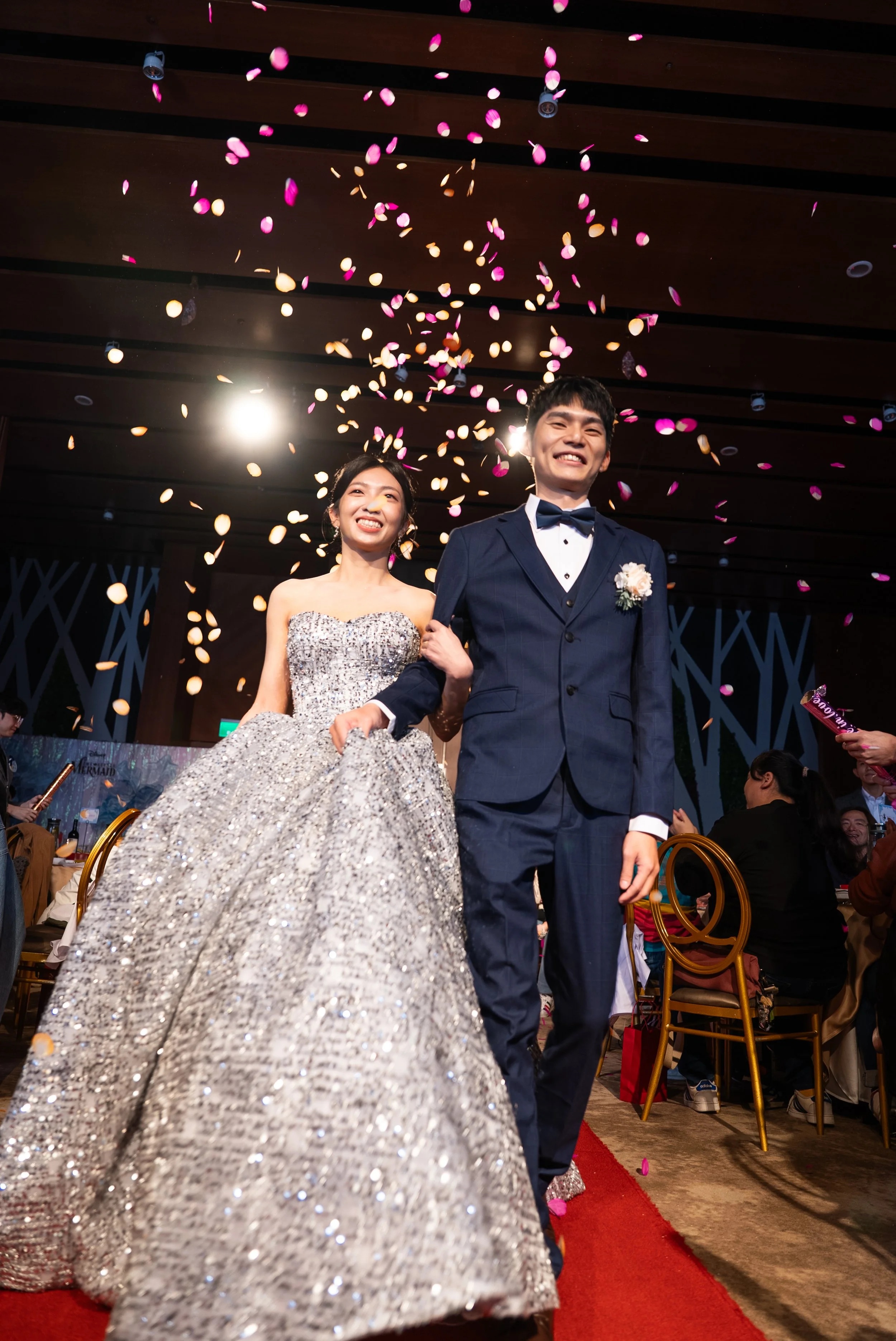 A bride and groom smiling as they walk down the aisle with confetti falling around them at their wedding reception.