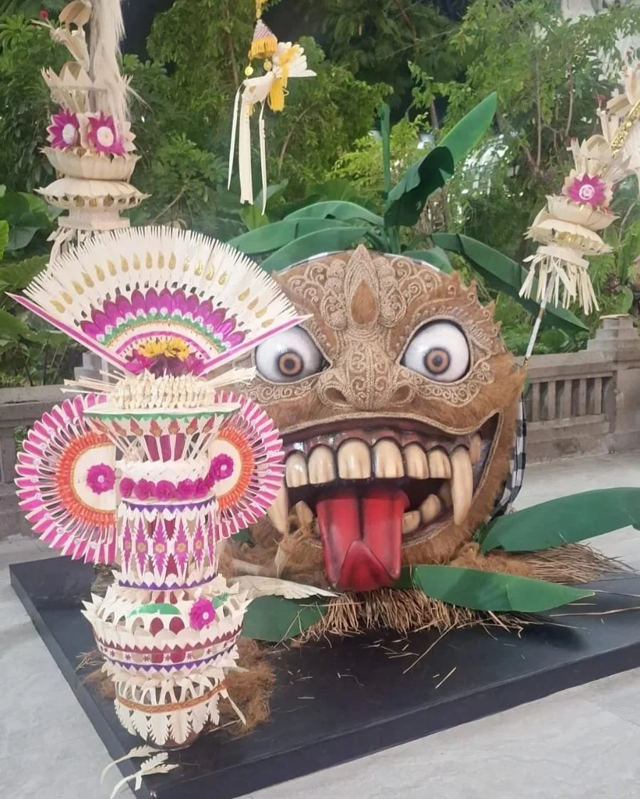 Decorative display featuring a large traditional carved monster face with bulging eyes, wide open mouth with large teeth and a long tongue, surrounded by green leaves. In front, colorful intricately designed tiered paper lanterns and ornaments with pink, white, and purple patterns. The setup is outdoors with a wooden fence and leafy plants in the background.