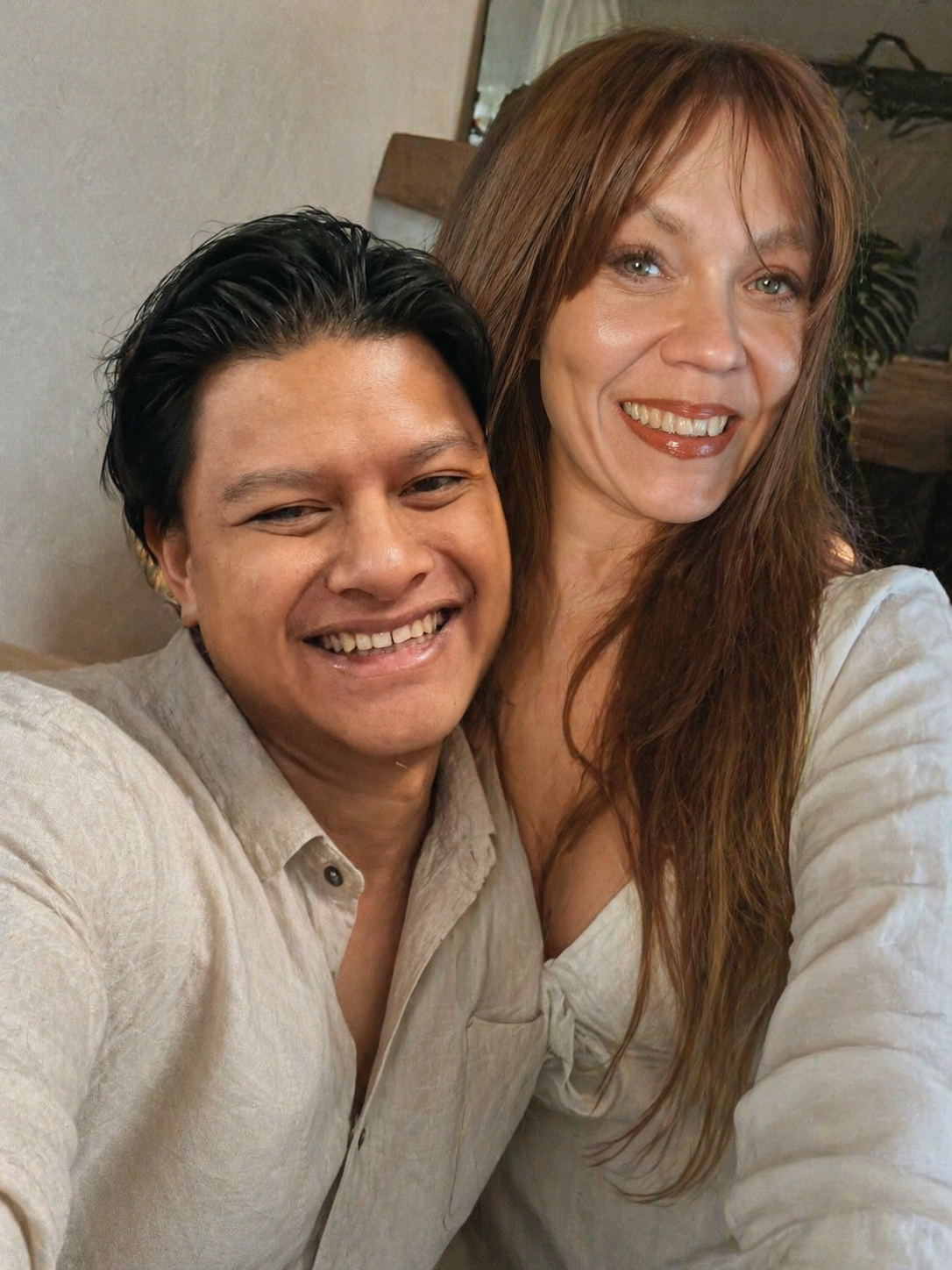 A smiling balinese man and a Finnish woman taking a selfie together in Bali. The man has short dark hair and is wearing a light-colored shirt, while the woman has long, wavy reddish-brown hair and is wearing a white top.