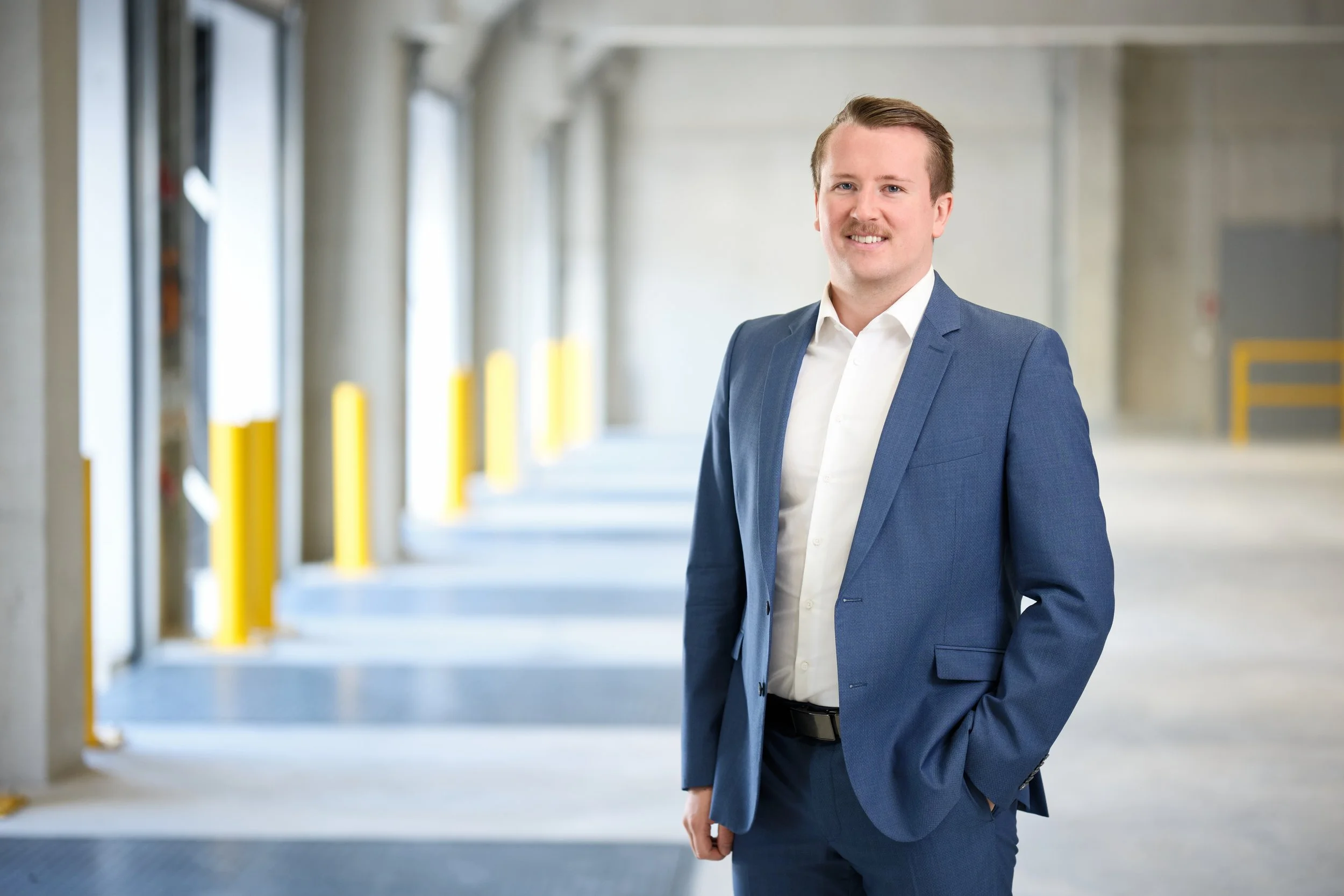 Ein Mann in einem blauen Anzug steht in einer Parkgarage und lächelt in die Kamera.