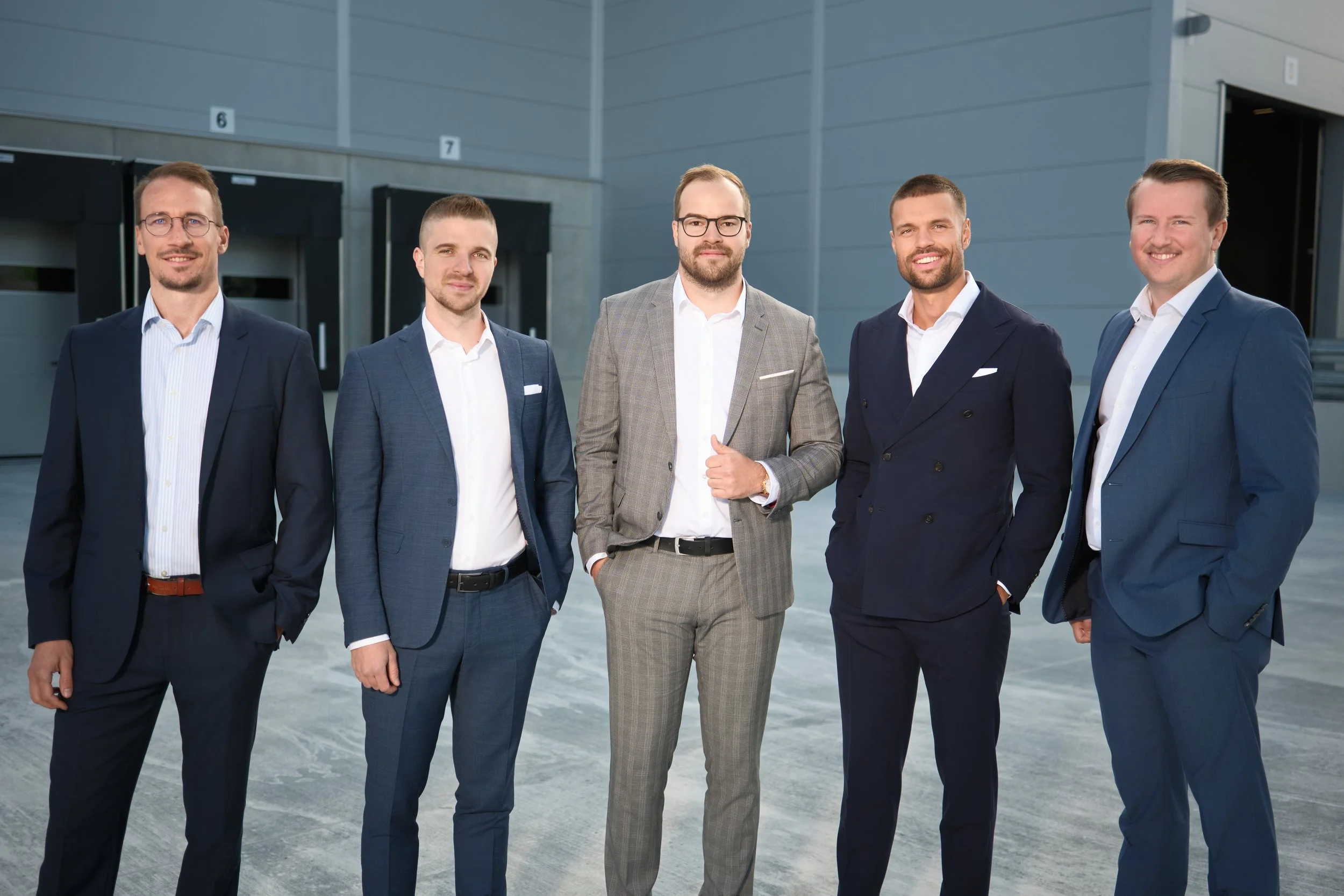 Sechs Männer in Businessanzügen stehen vor einem modernen Gebäude.