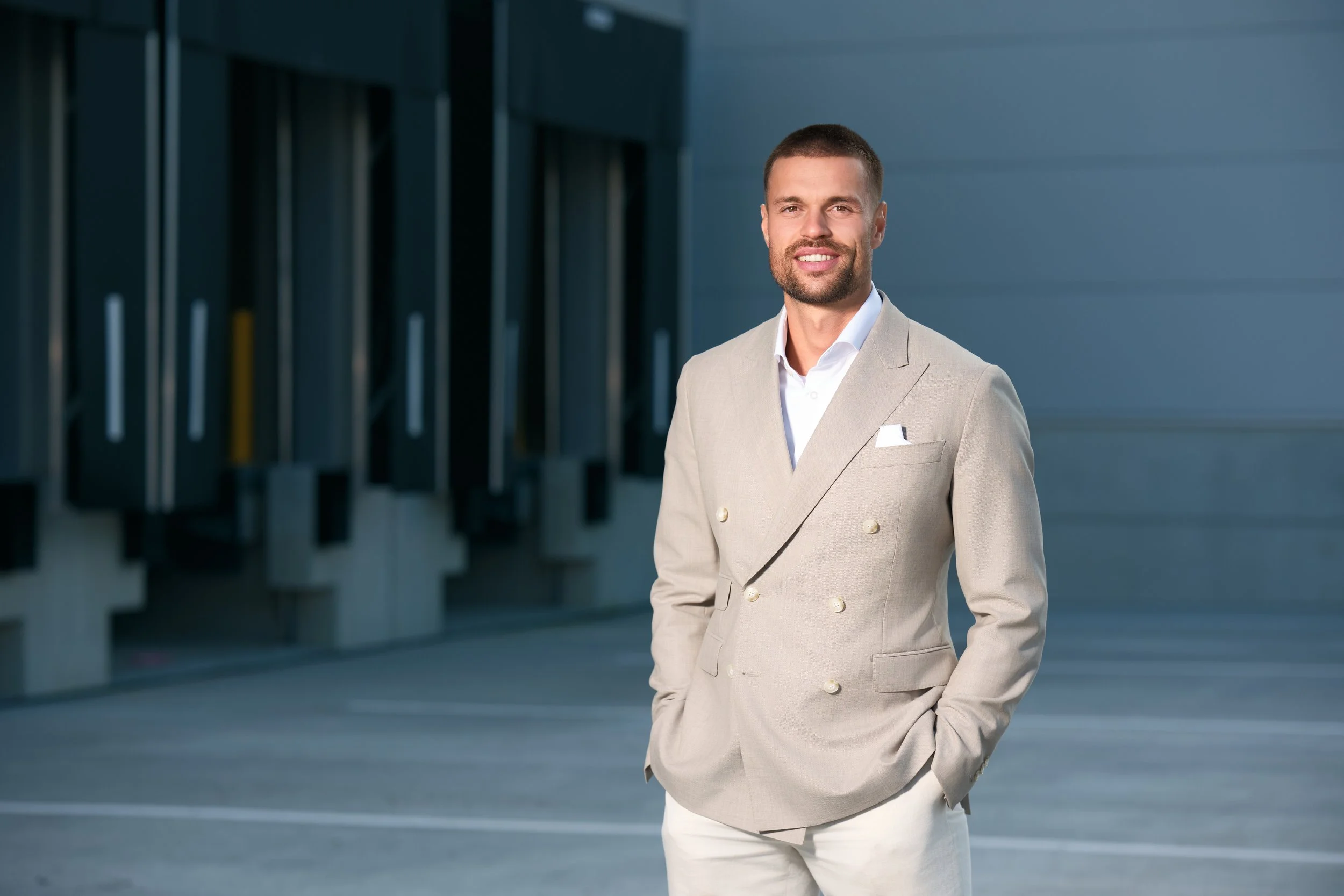 Mann in beigem Anzug mit weißen Hosen steht auf einer Parkfläche vor einem modernen Gebäude.