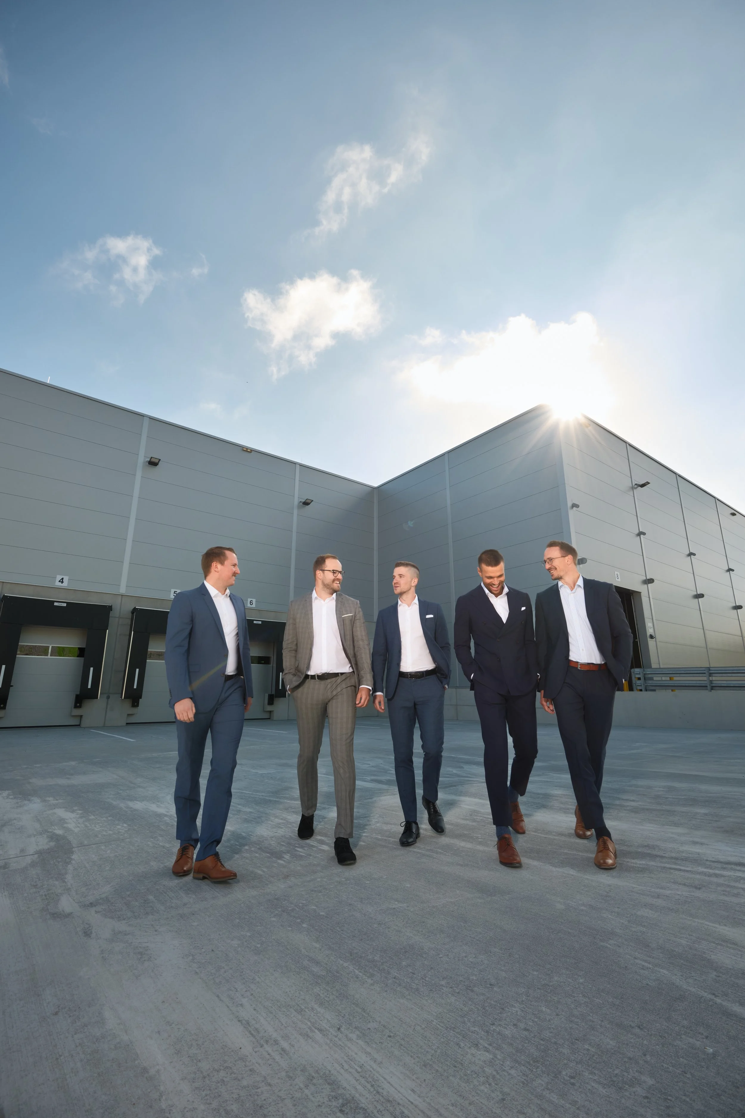 Eine Gruppe von sechs Männern in Anzügen, die auf einer Parkfläche vor einem modernen Gebäude spazieren.