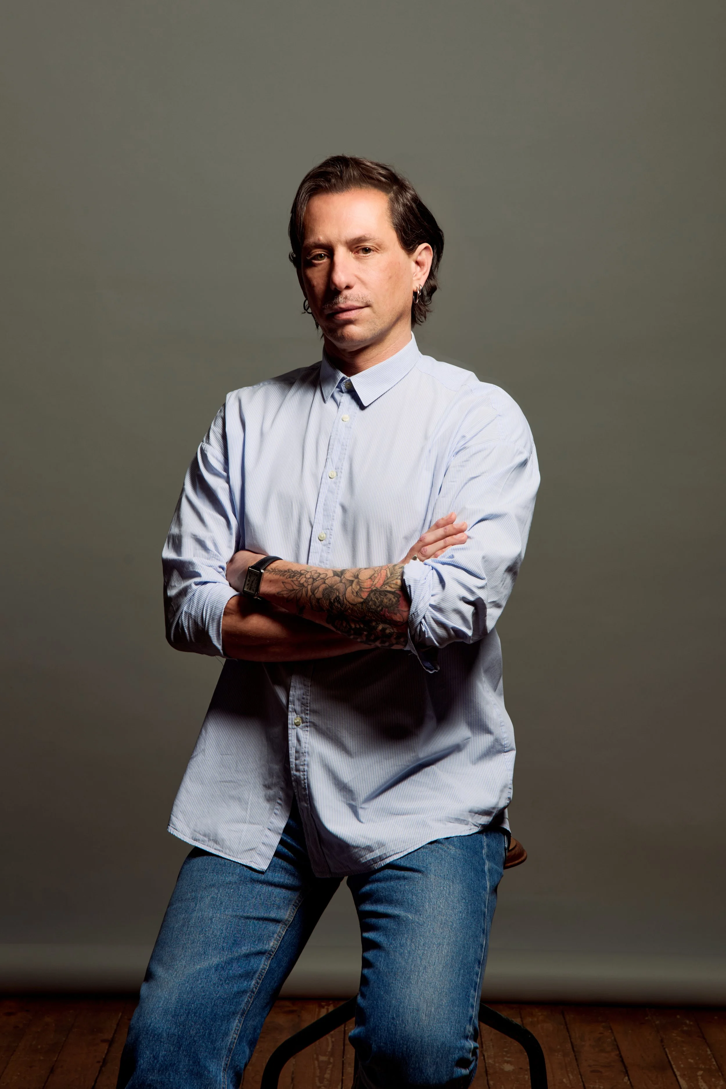 Un homme aux cheveux foncés, portant une chemise à manches longues et un jean, pose avec les bras croisés, un tatouage visible sur son bras droit, sur un fond gris