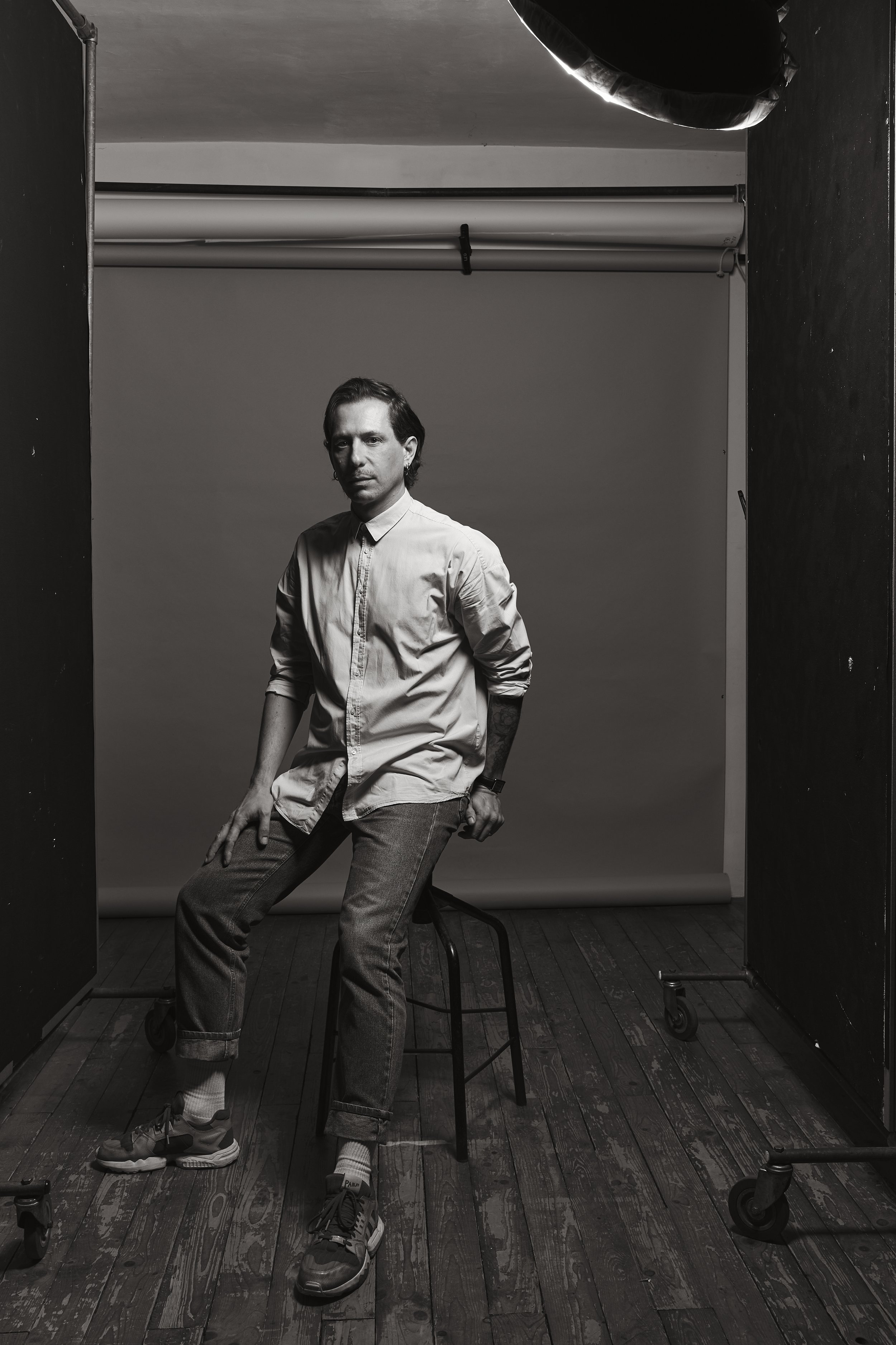Un homme assis sur une chaise en studio de photographie, noir et blanc, avec un décor simple et un éclairage professionnel.