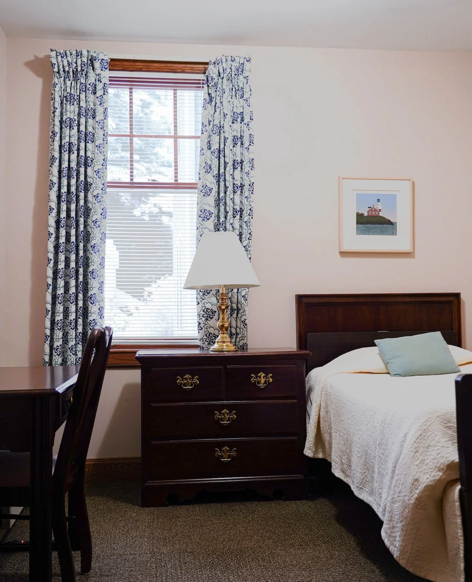 A bedroom in Lockwood with window, white curtains with blue floral pattern, wooden window frame, dresser with a lamp, bed with a white quilt, and framed picture of a lighthouse hanging above the bed.