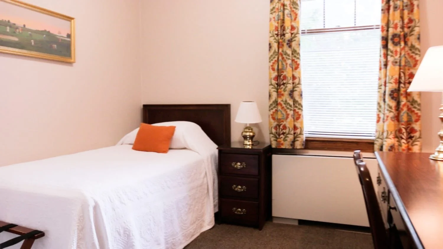 A bedroom in Lockwood with a single bed, a wooden nightstand with a gold lamp, orange pillow, window with patterned floral curtains, and a wooden desk with a chair.