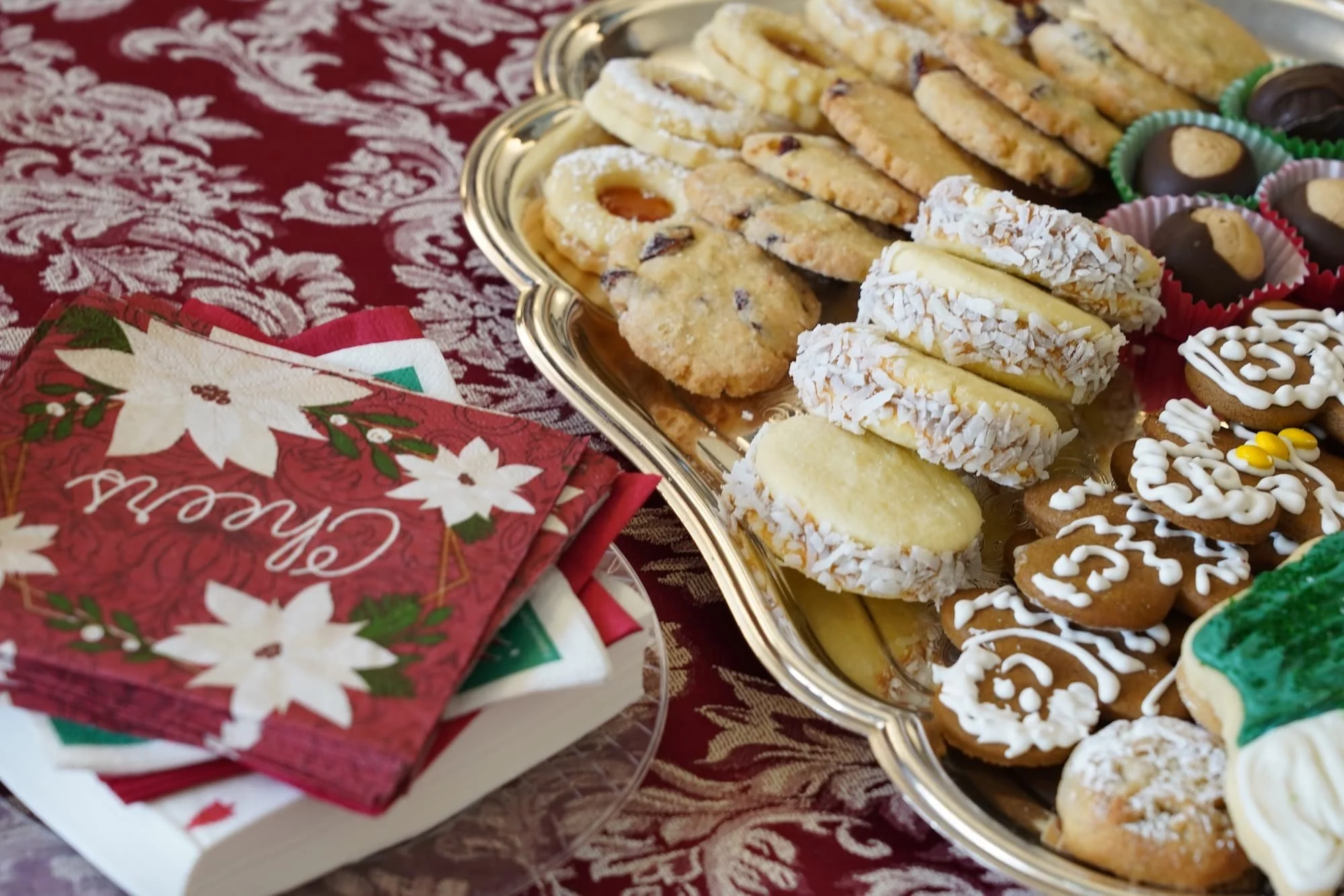 A silver tray filled with assorted Christmas cookies including sugar cookies with coconut, chocolate-covered cookies, and decorated gingerbread cookies is placed on a red holiday tablecloth. A box of Christmas napkins with poinsettia and holiday designs is next to the tray.