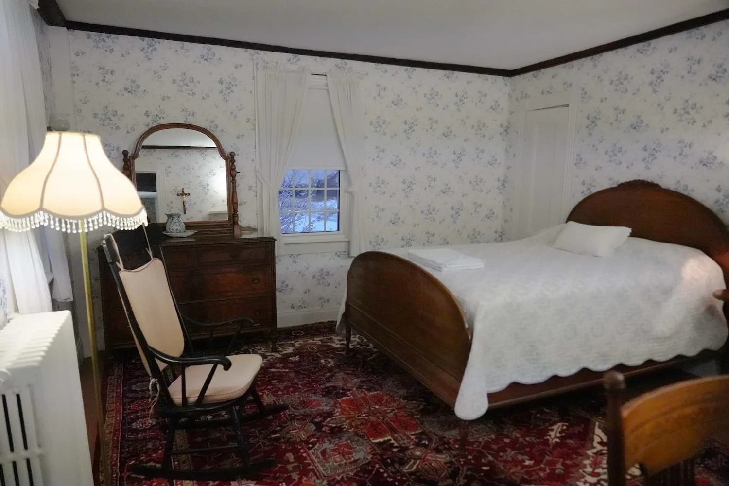 A main house vintage bedroom with a wooden bed covered in white bedding, a wooden dresser with a mirror, a chair, a lamp, a window with curtains, and floral wallpaper.