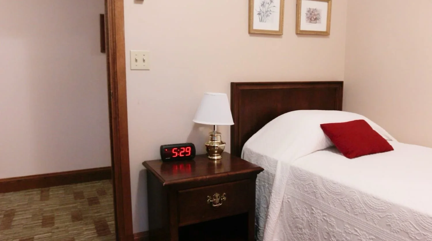 A bedroom in Lockwood with a wooden nightstand, white lamp, digital clock showing 5:29, bed with white quilt and red pillow, and framed botanical artwork on the wall.