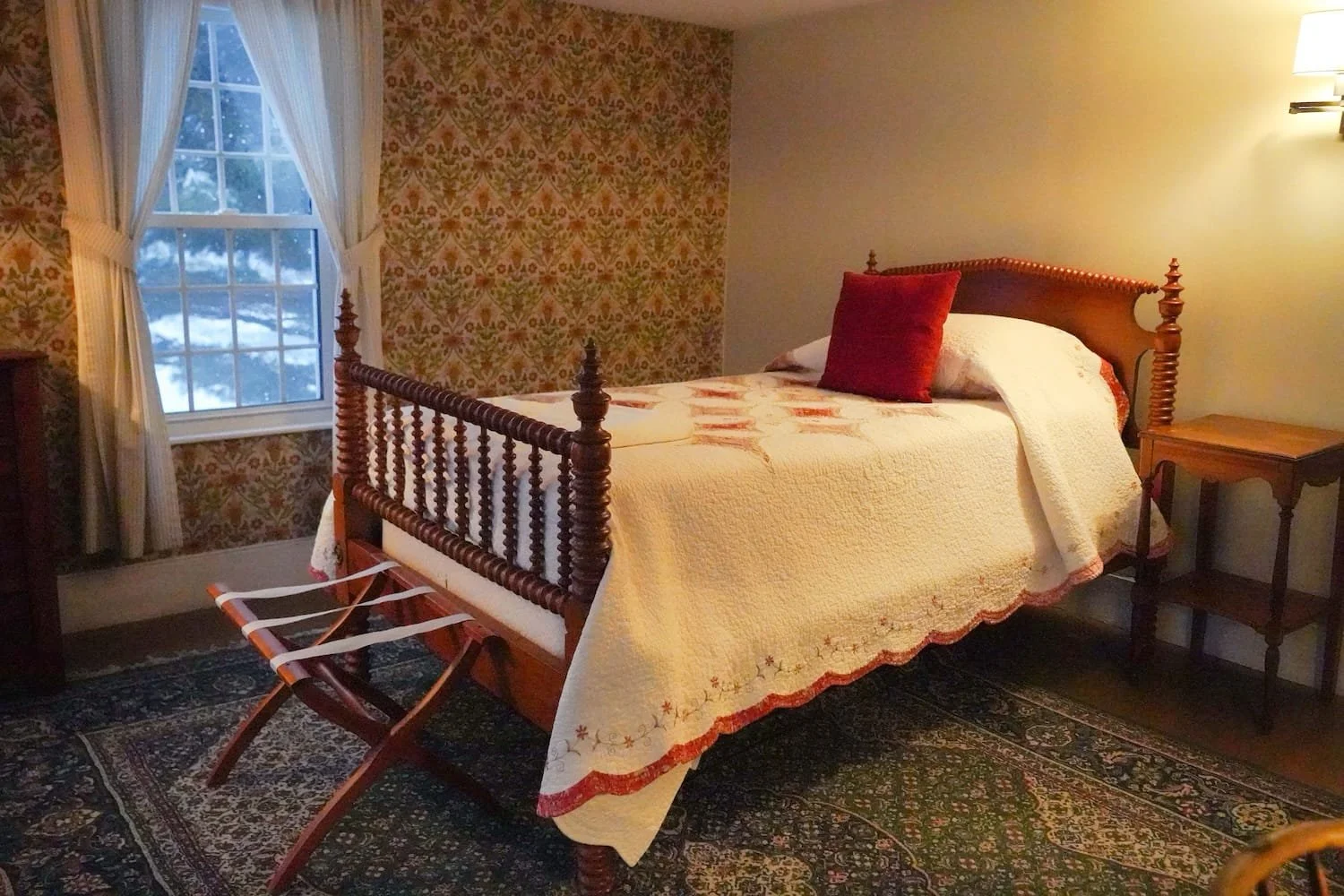 A main house bedroom with a wooden bed covered by a quilt and a red pillow, beside a window with white curtains, a side table, and a patterned rug.