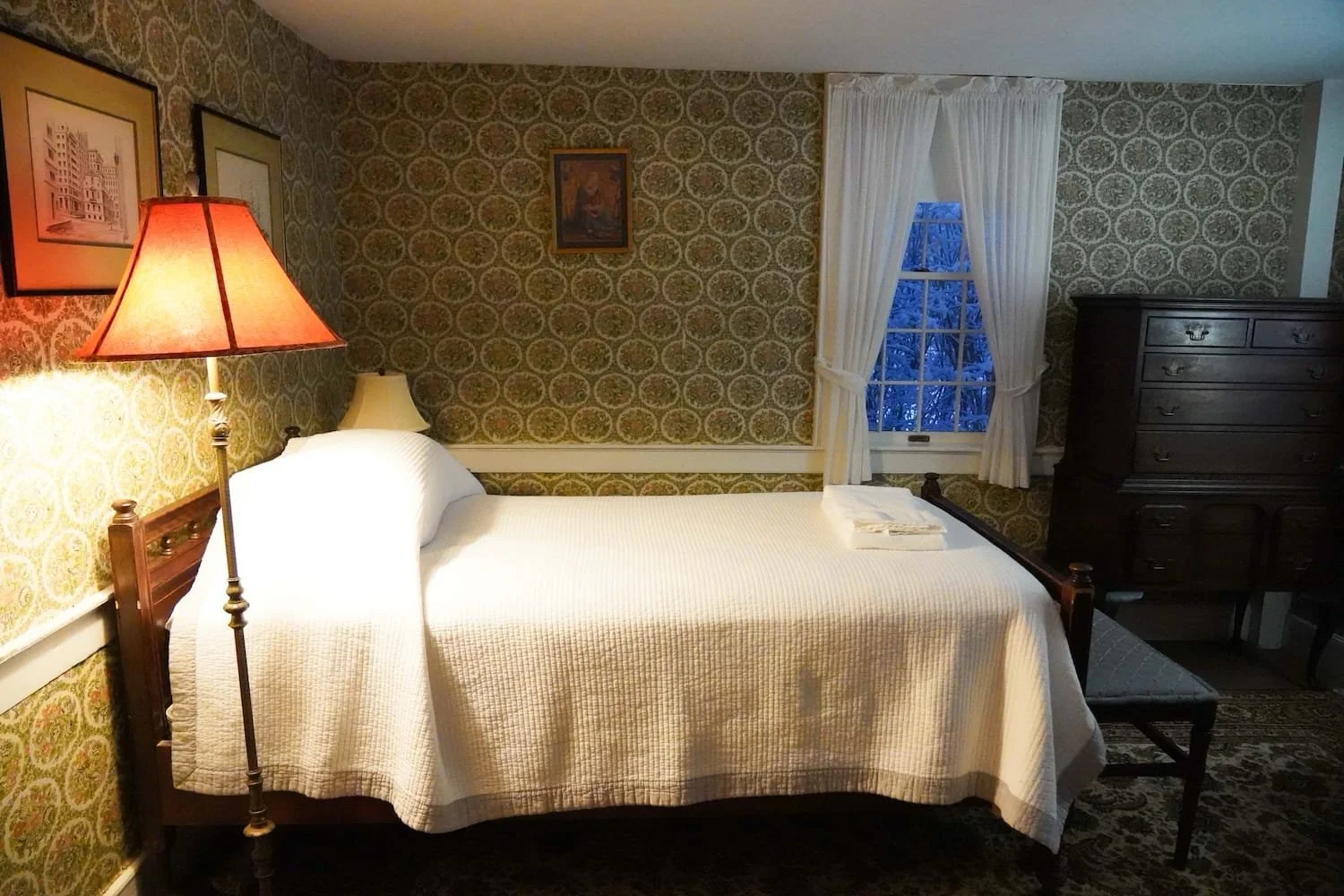 A main house bedroom with a bed covered in a white quilt, a wooden bench at the foot of the bed, a tall dresser to the right, a window with white curtains, a lamp with a pink shade on a side table, and framed pictures on the wallpapered walls.