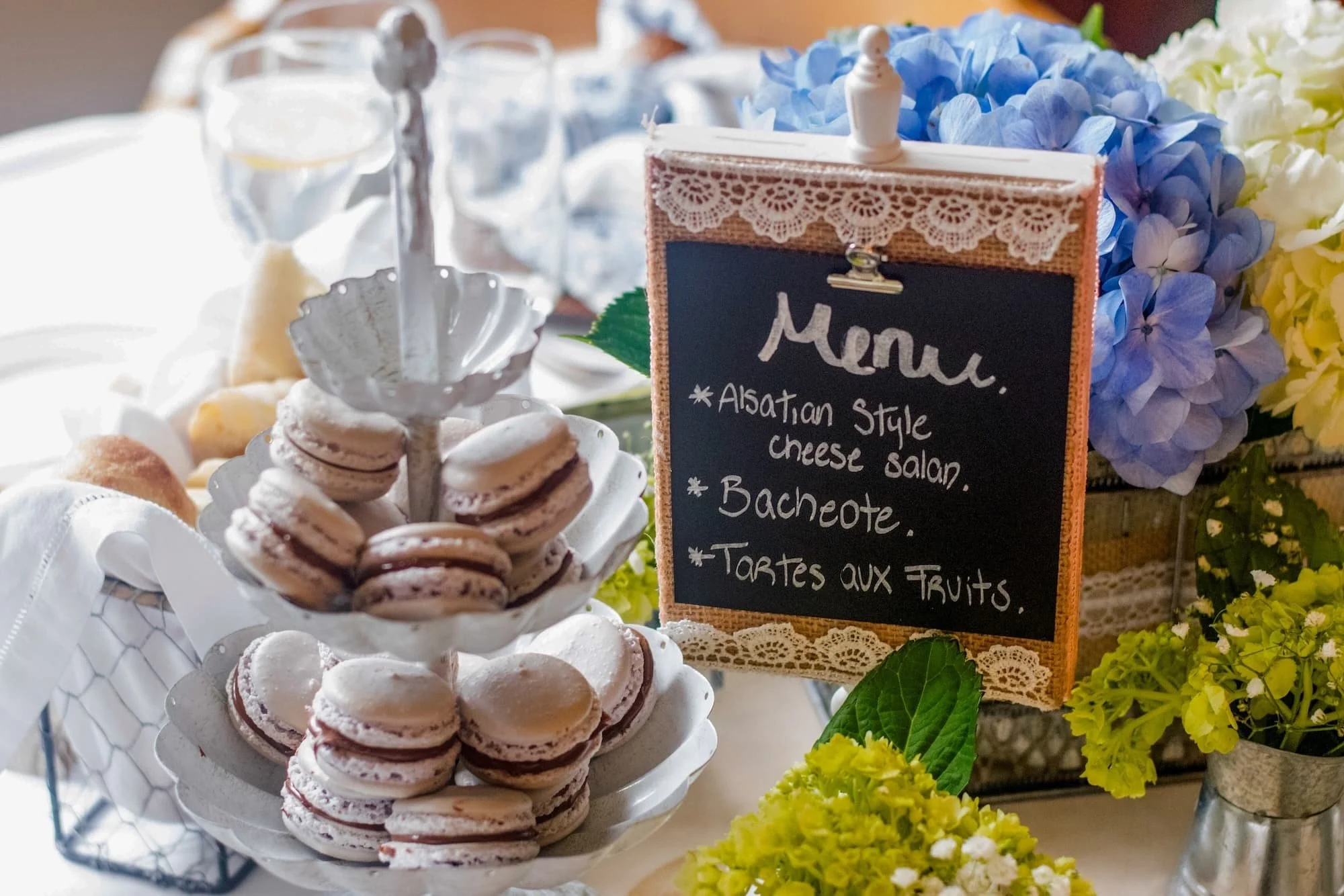 A tiered plate of assorted macarons on a white doily, with a menu card beside it listing Albanian-style cheese salad, baked item, and fruit tartes, surrounded by colorful hydrangea flowers and decorative greenery.