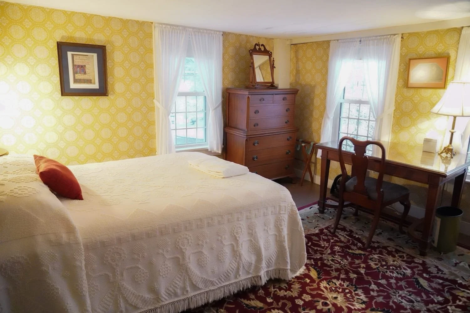 A main house cozy bedroom with a bed with a white bedspread and a red pillow, yellow patterned wallpaper, two windows with sheer white curtains, a wooden dresser with a mirror, a desk with a wooden chair, a lamp, and a decorative red and beige rug.