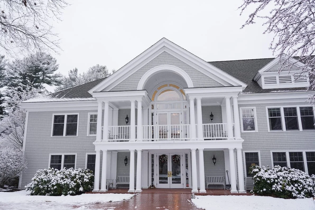 Lockwood building with a portico entrance, old style black lanterns, and snow-covered bushes in front of a snow-dusted landscape under a cloudy sky.