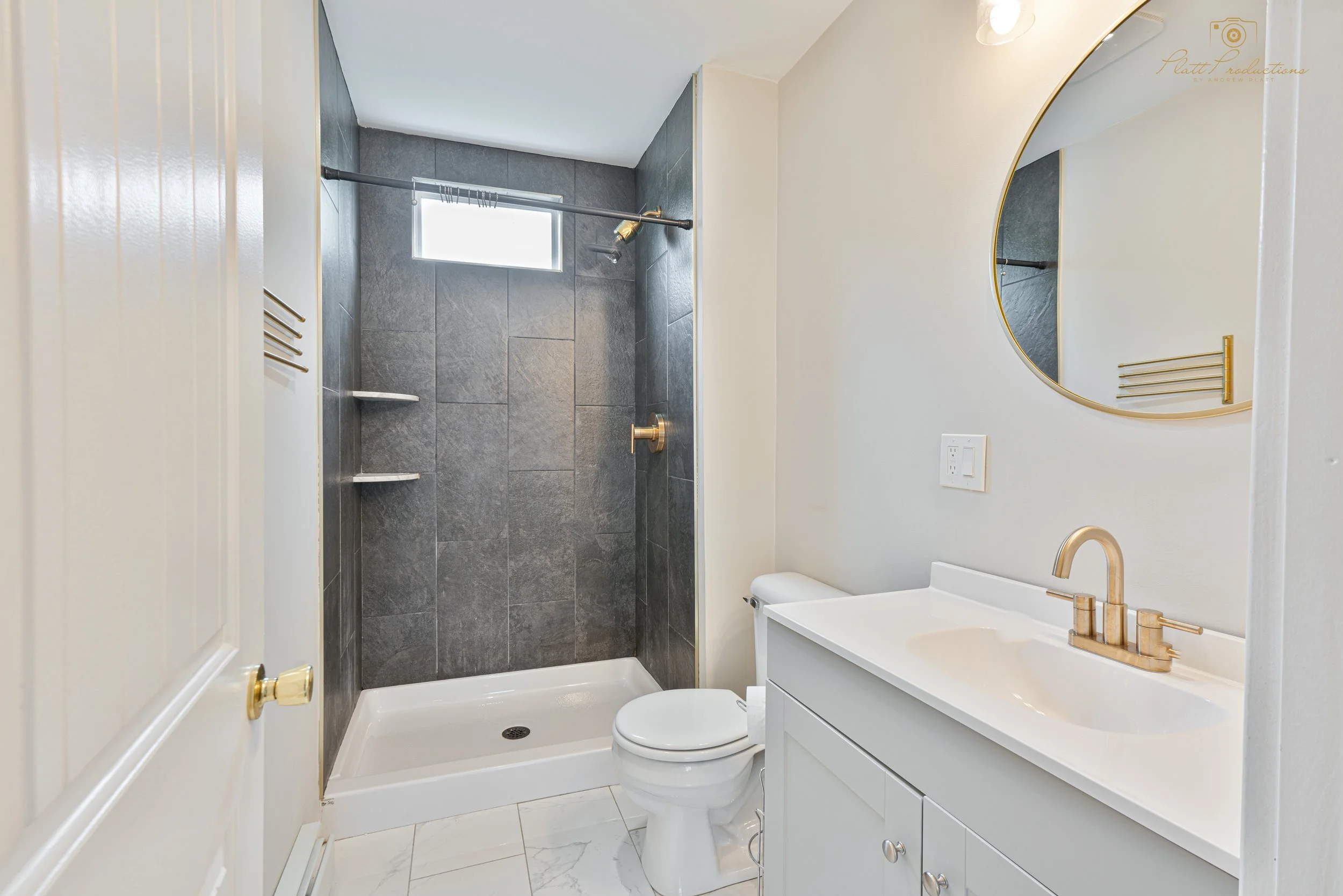 Bathroom with shower area featuring dark gray tiled walls, a small high window, a white toilet, white vanity with a sink, a round mirror, and gold fixtures.