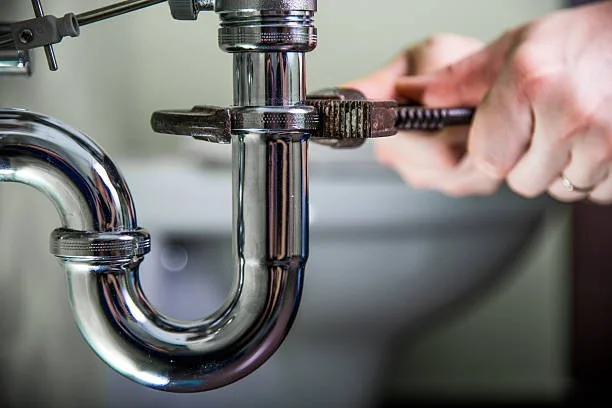 A person repairing or installing a metal pipe under a sink using a pipe wrench.