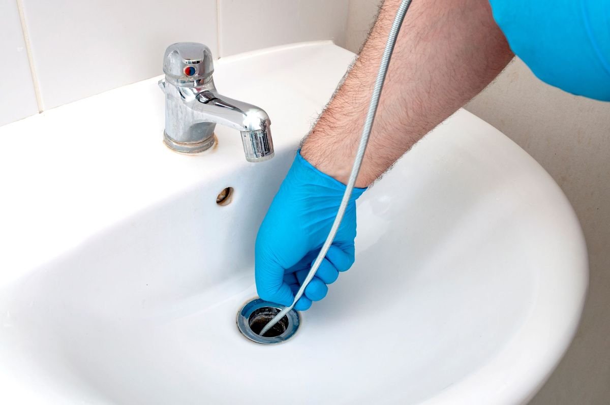 A person wearing blue gloves is cleaning a bathroom sink drain with a long tool.