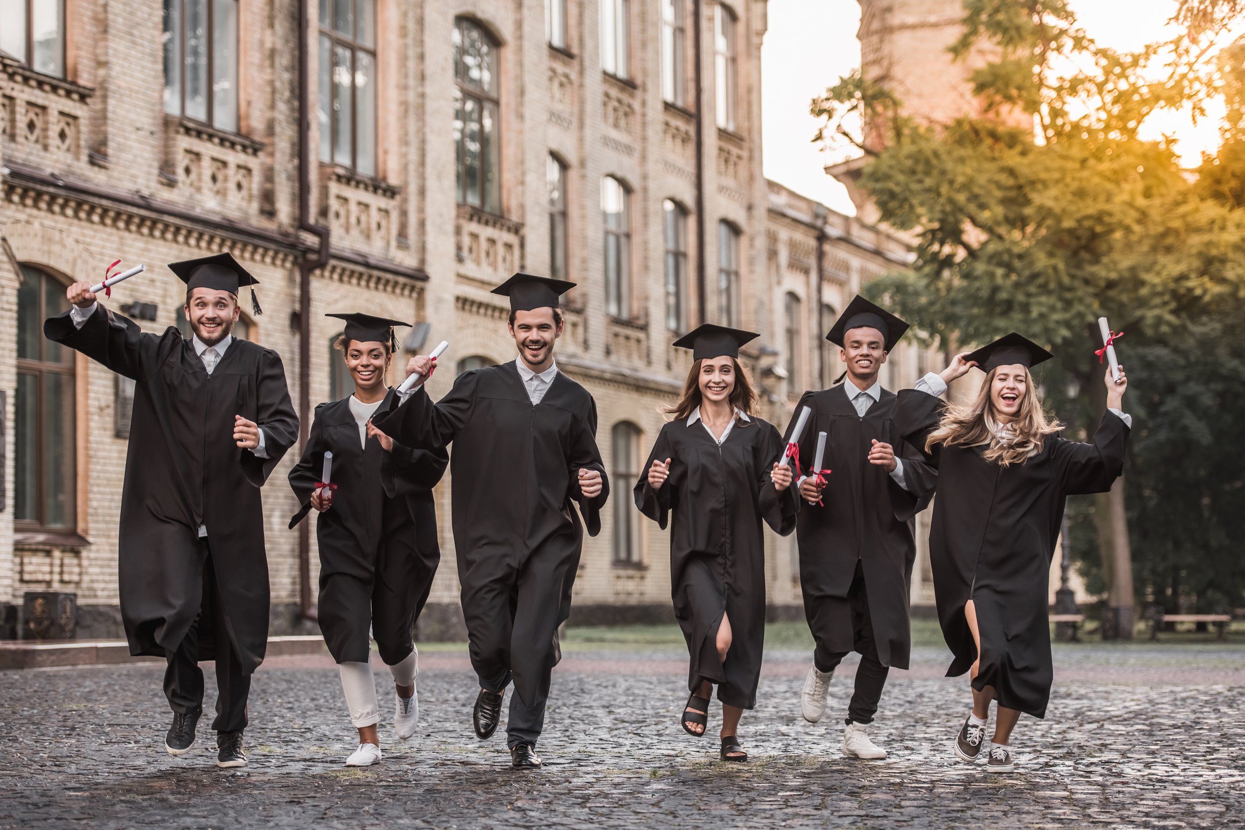 A group of six diverse college graduates in caps and gowns celebrating outdoors, holding diplomas, and running on a cobblestone path with a historic building and trees in the background during sunset.
