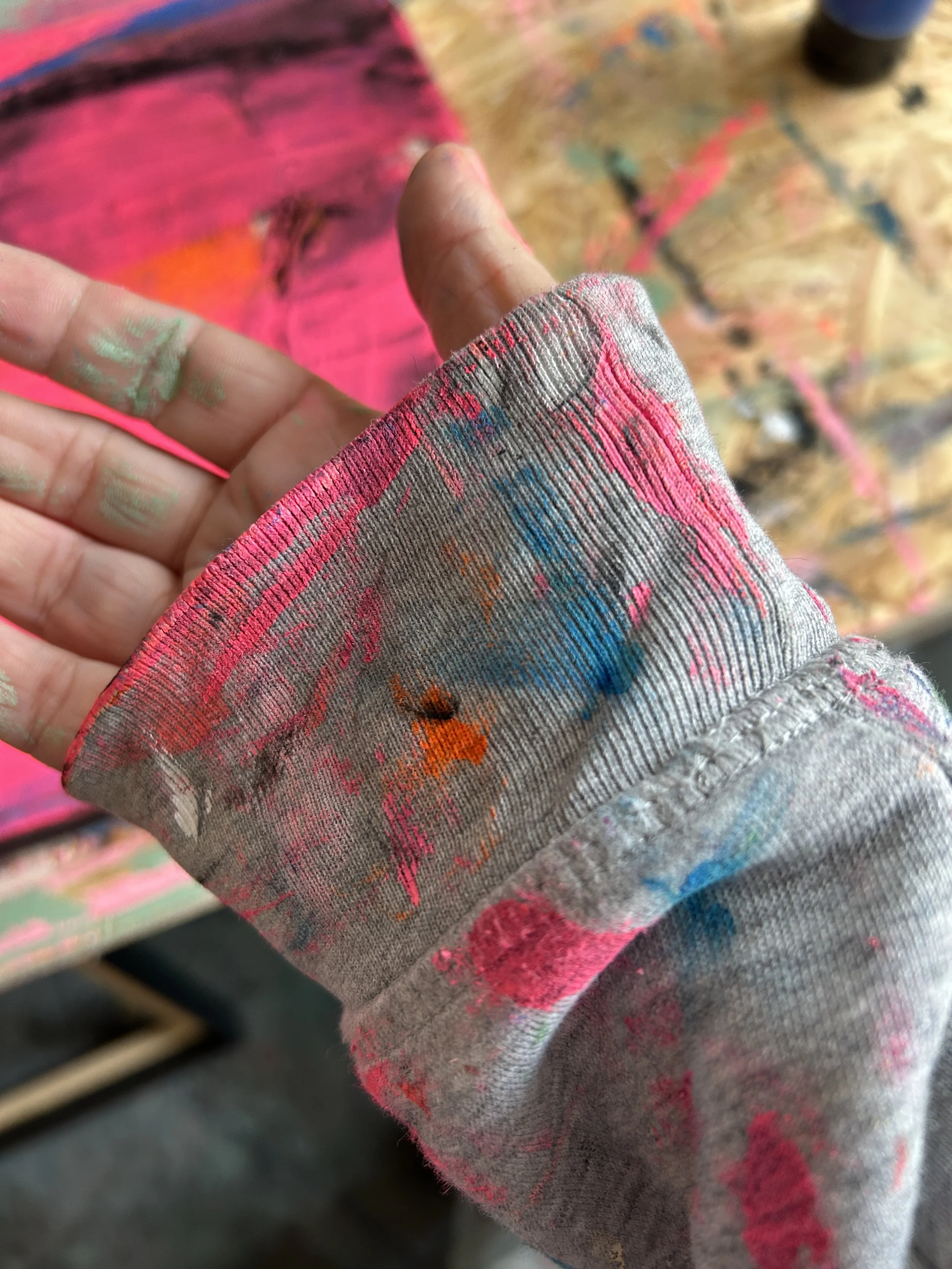 Eine Hand, die einen fusselfreien, mit Farbspritzern bedeckten Stoff hält, auf einem bunten, klebrigen Arbeitsbereich mit Farbspritzern im Hintergrund.