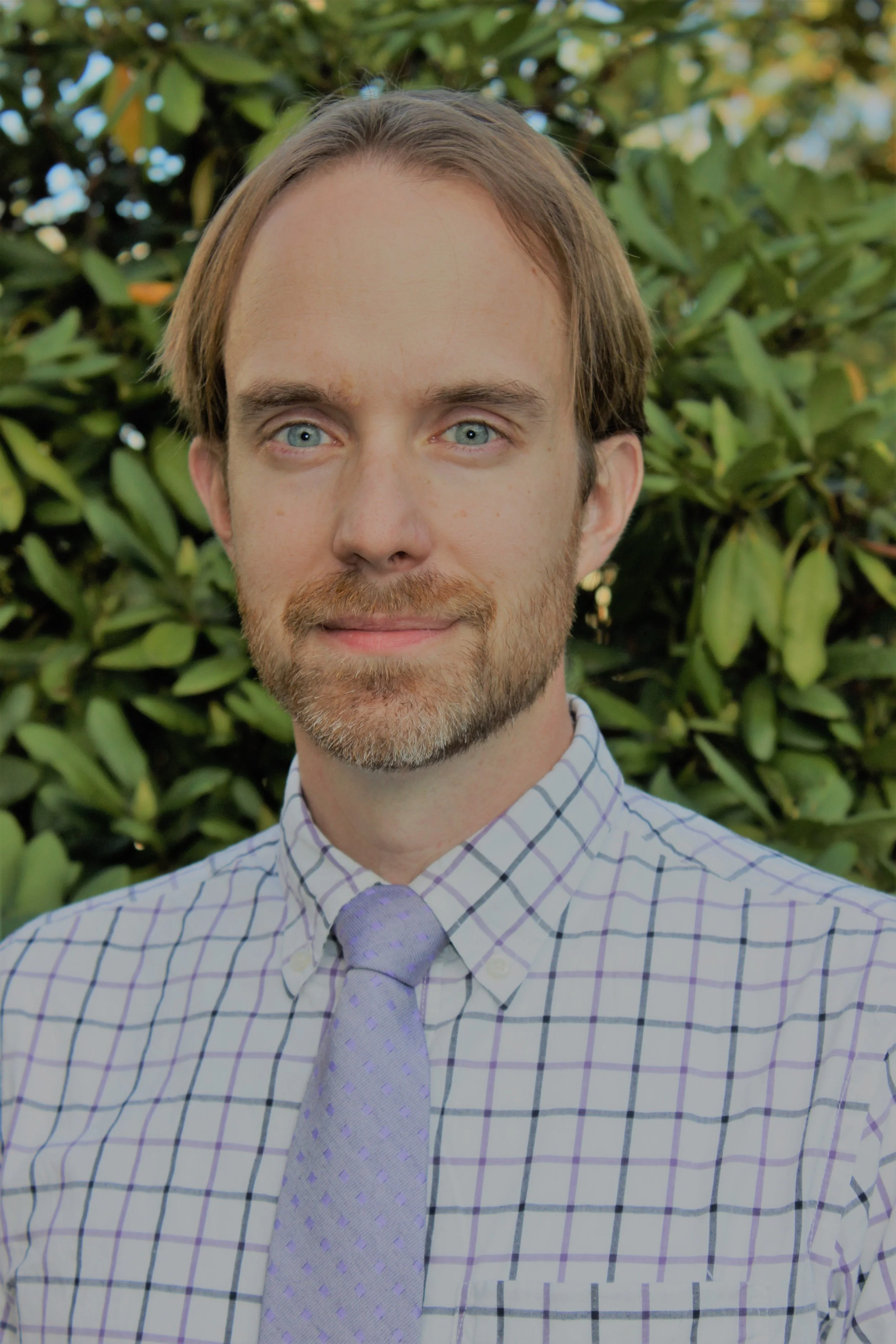 A man with blue eyes, light brown hair, and a beard, wearing a checkered shirt and a lavender tie, standing outdoors in front of green shrubbery.