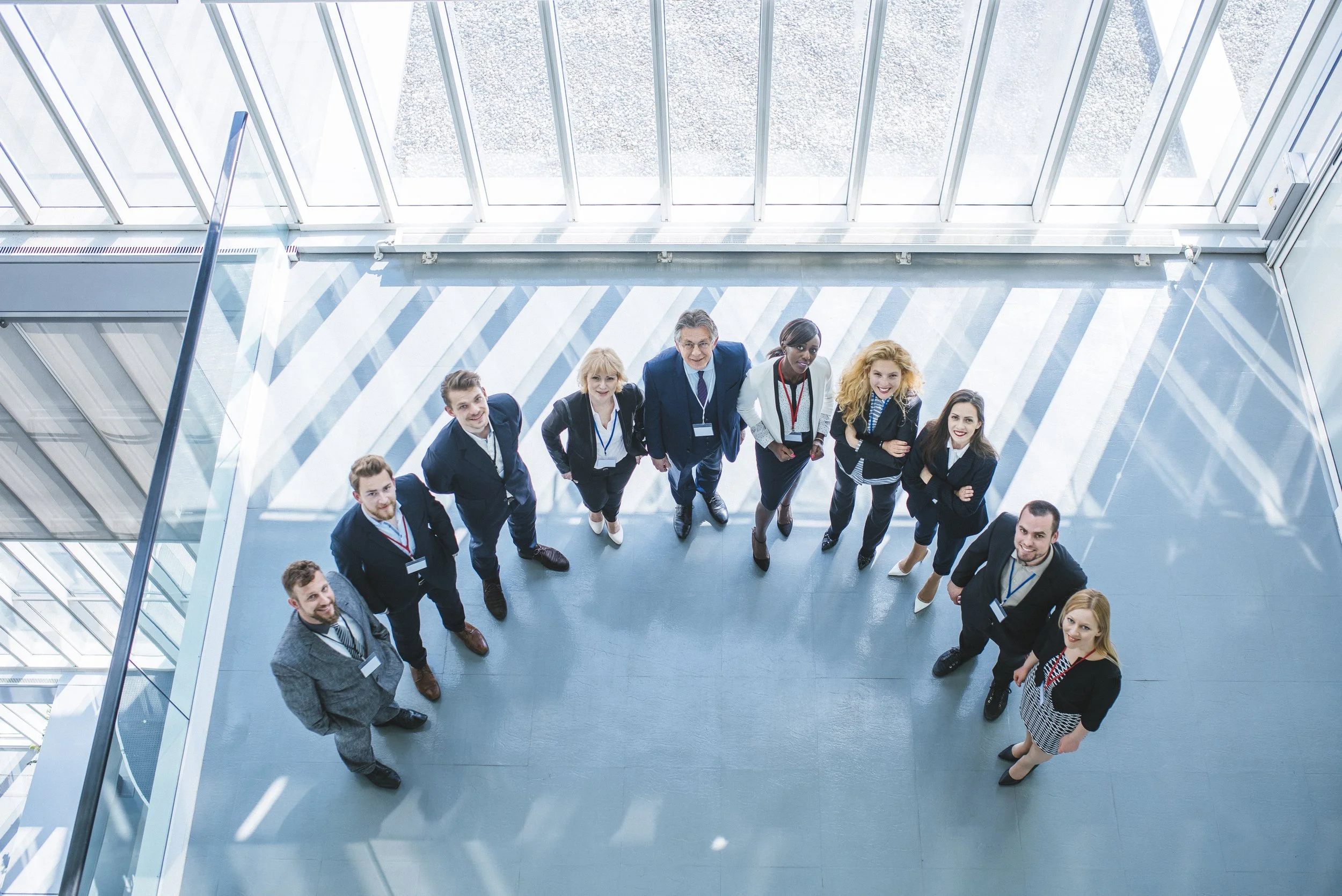 A group of eleven diverse business professionals looking up at the camera in a modern office building with large glass windows. Recruiter.