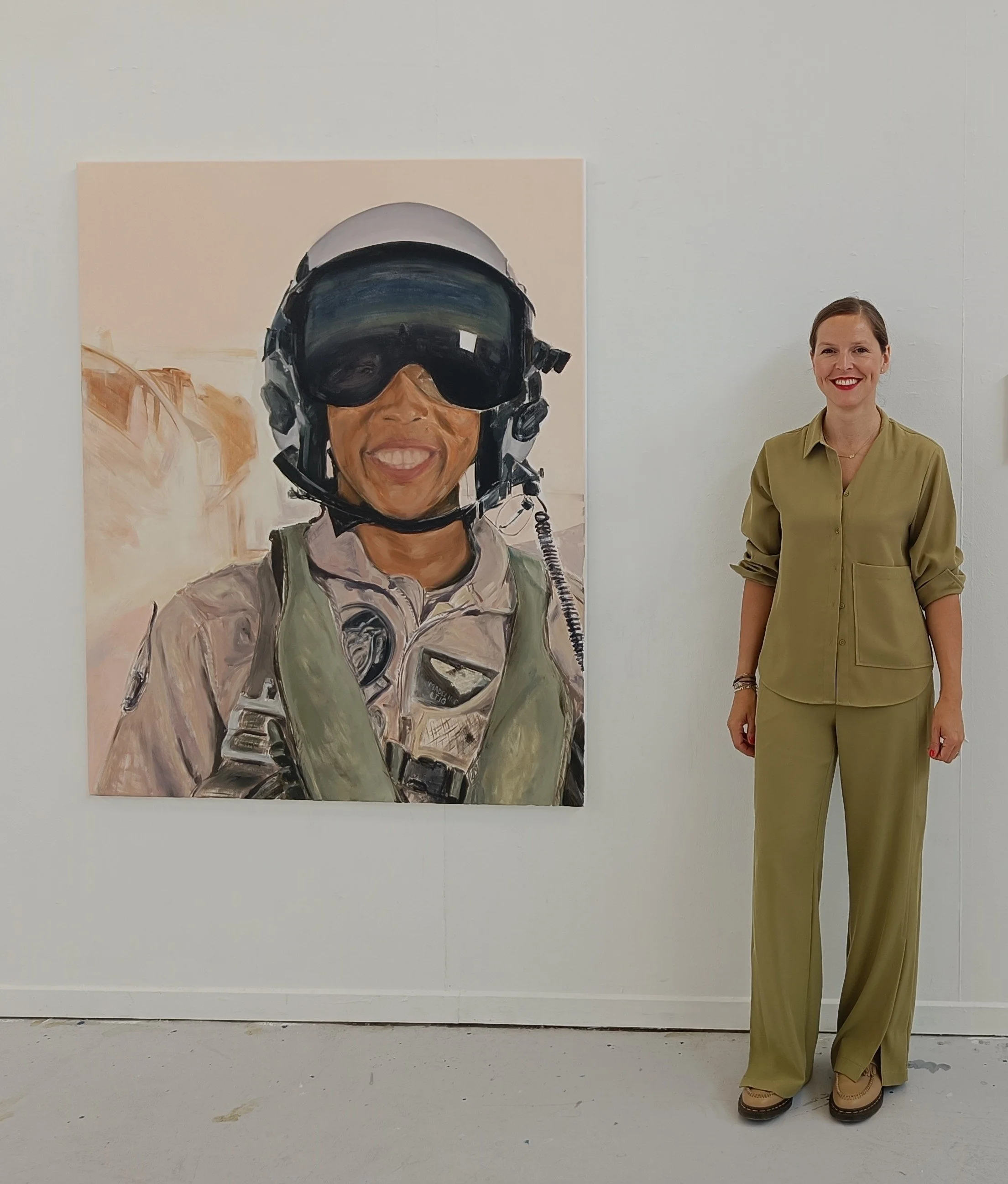 Artist Truus Van Bosstraeten in a green outfit standing beside her painting ‘Madeline Swegle' from the Hard Cover series. The large portrait depicts a female military pilot wearing a helmet and flight gear.