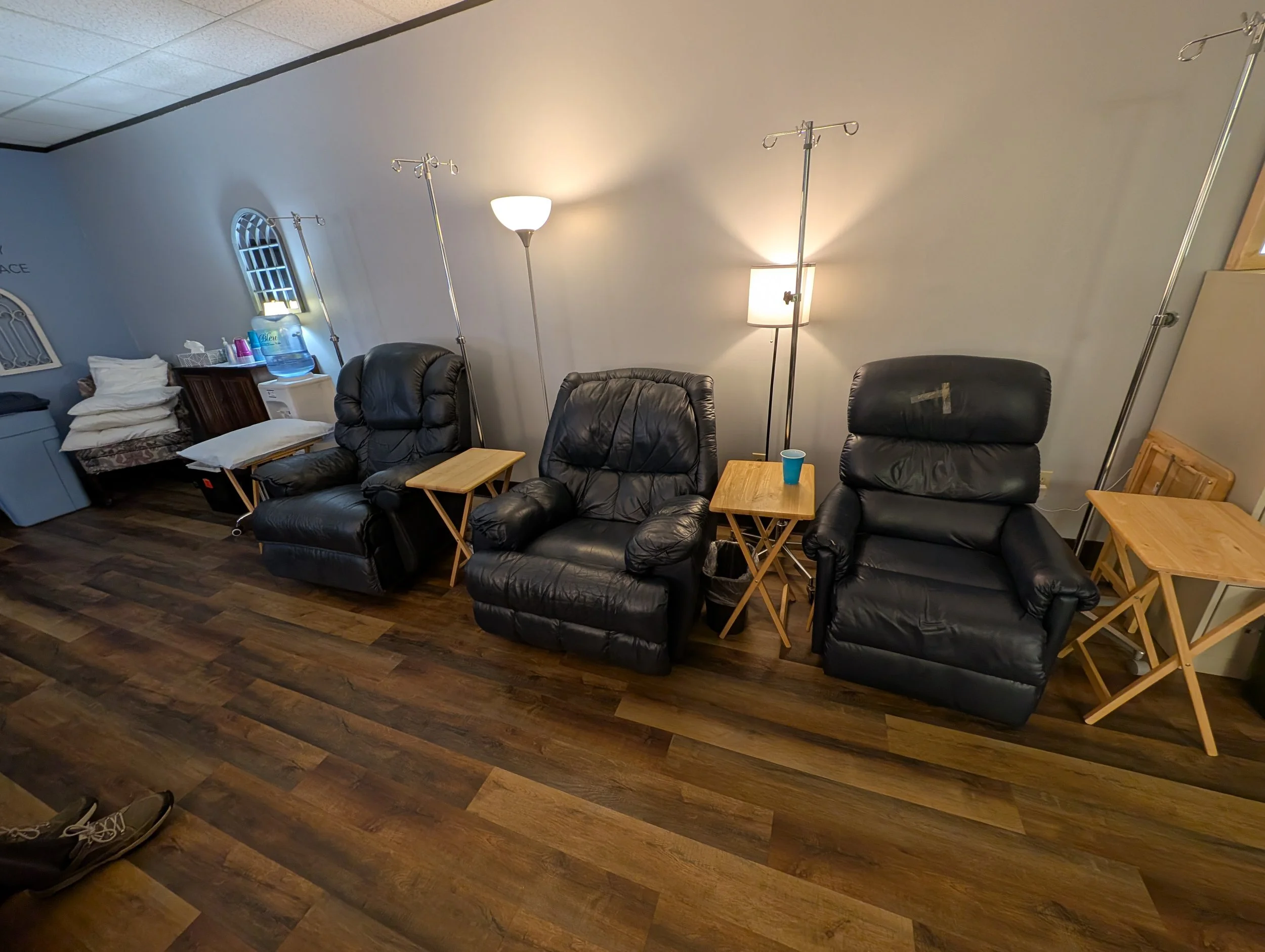Three black leather recliner chairs with small wooden side tables and various lamps in a room with wood flooring.