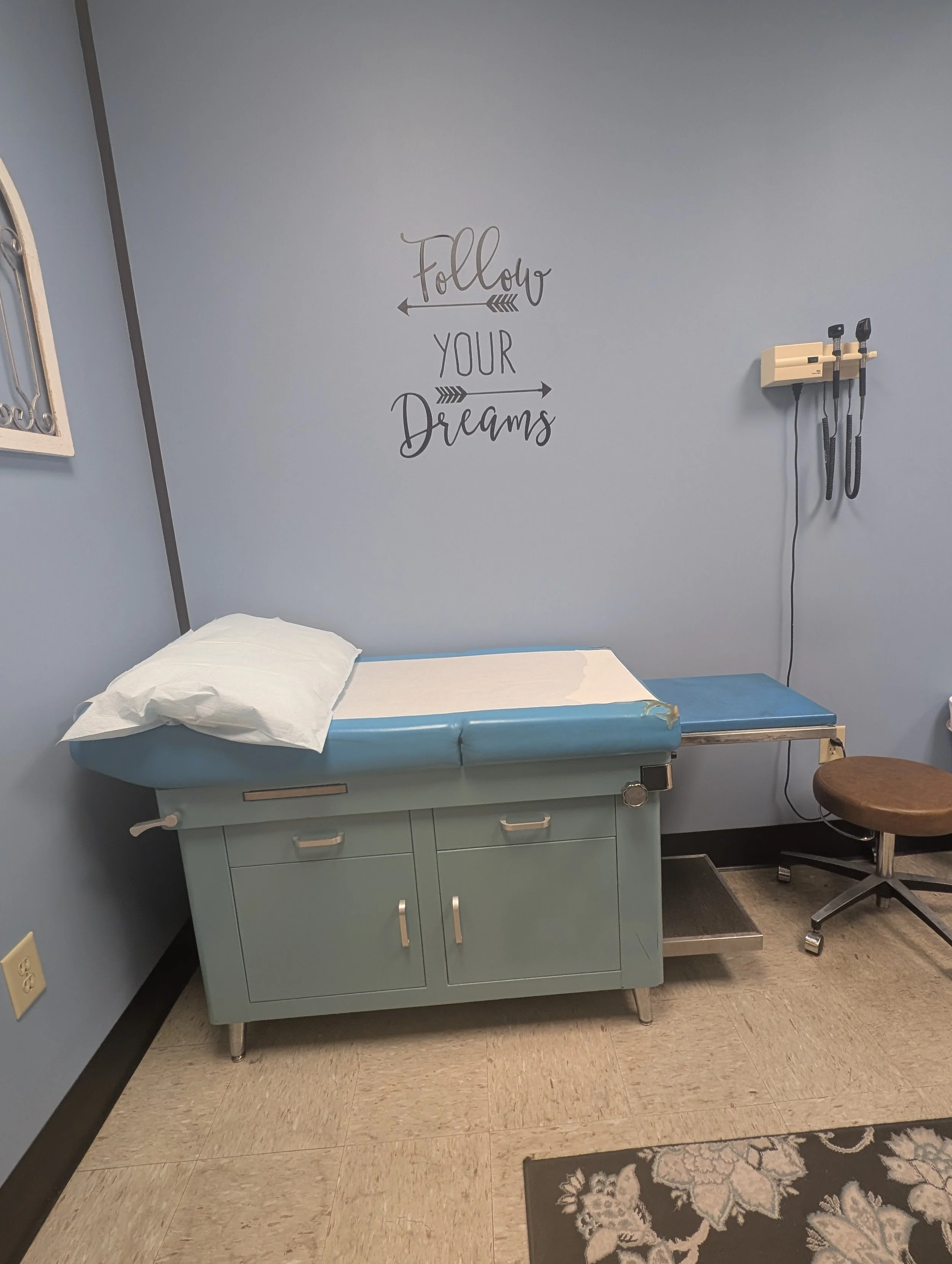 Medical examination room with a blue examination table, white pillow, wall decor with the phrase 'Follow Your Dreams', wall-mounted medical tools, stool, and a patterned rug.